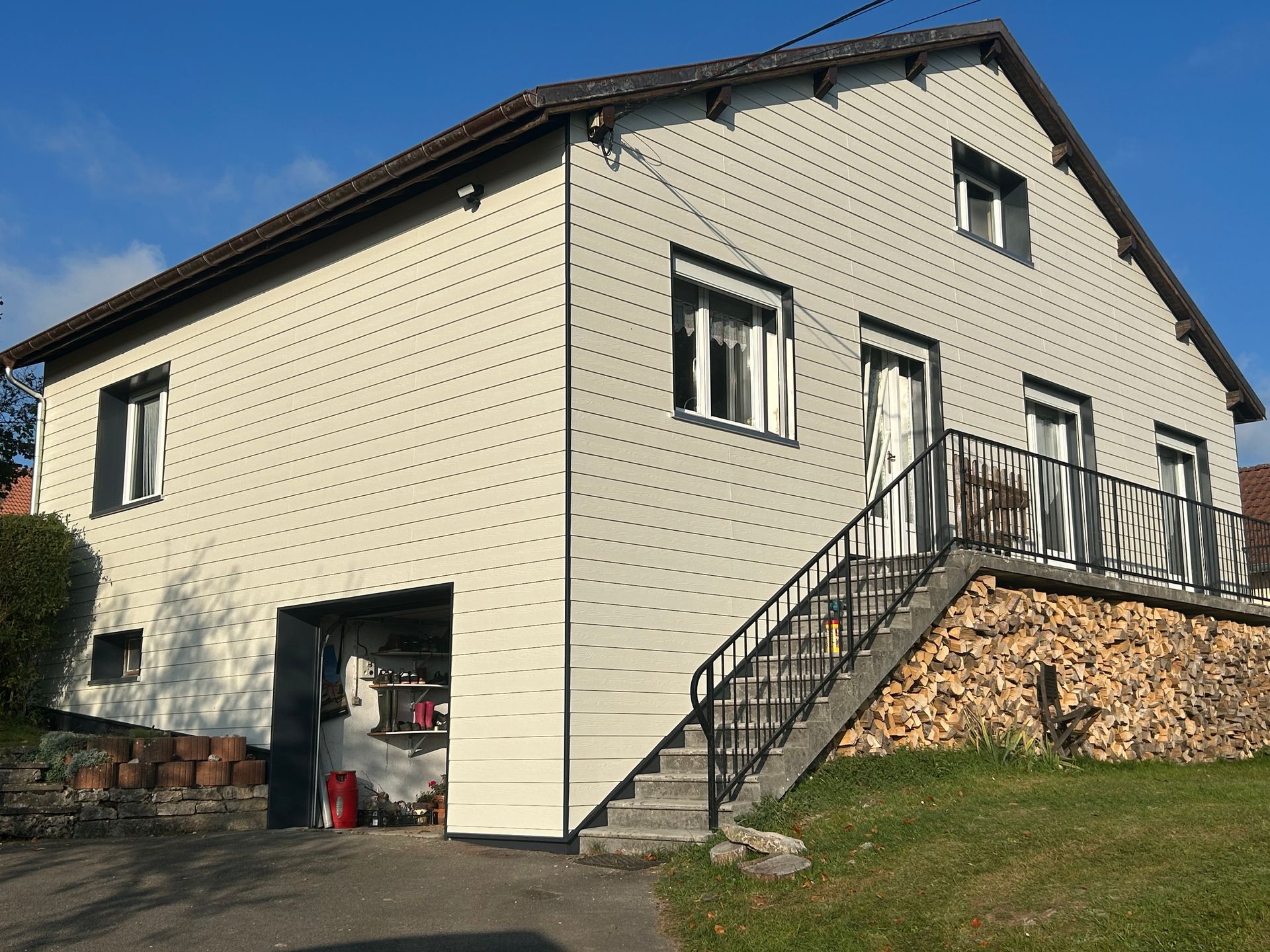 Une maison aux tons clairs avec un garage, un escalier extérieur menant à un balcon et un soubassement en pierre, sous un ciel dégagé.