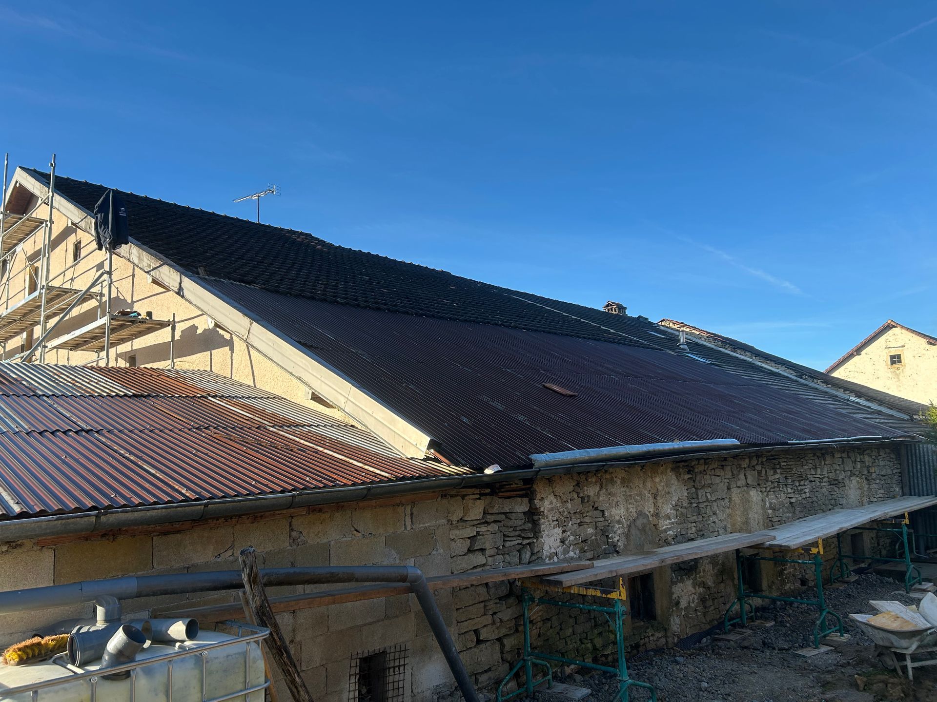 Un vieux bâtiment en pierre en construction, avec une toiture à moitié installée, composée de nouvelles tuiles noires et de panneaux bruns plus anciens.