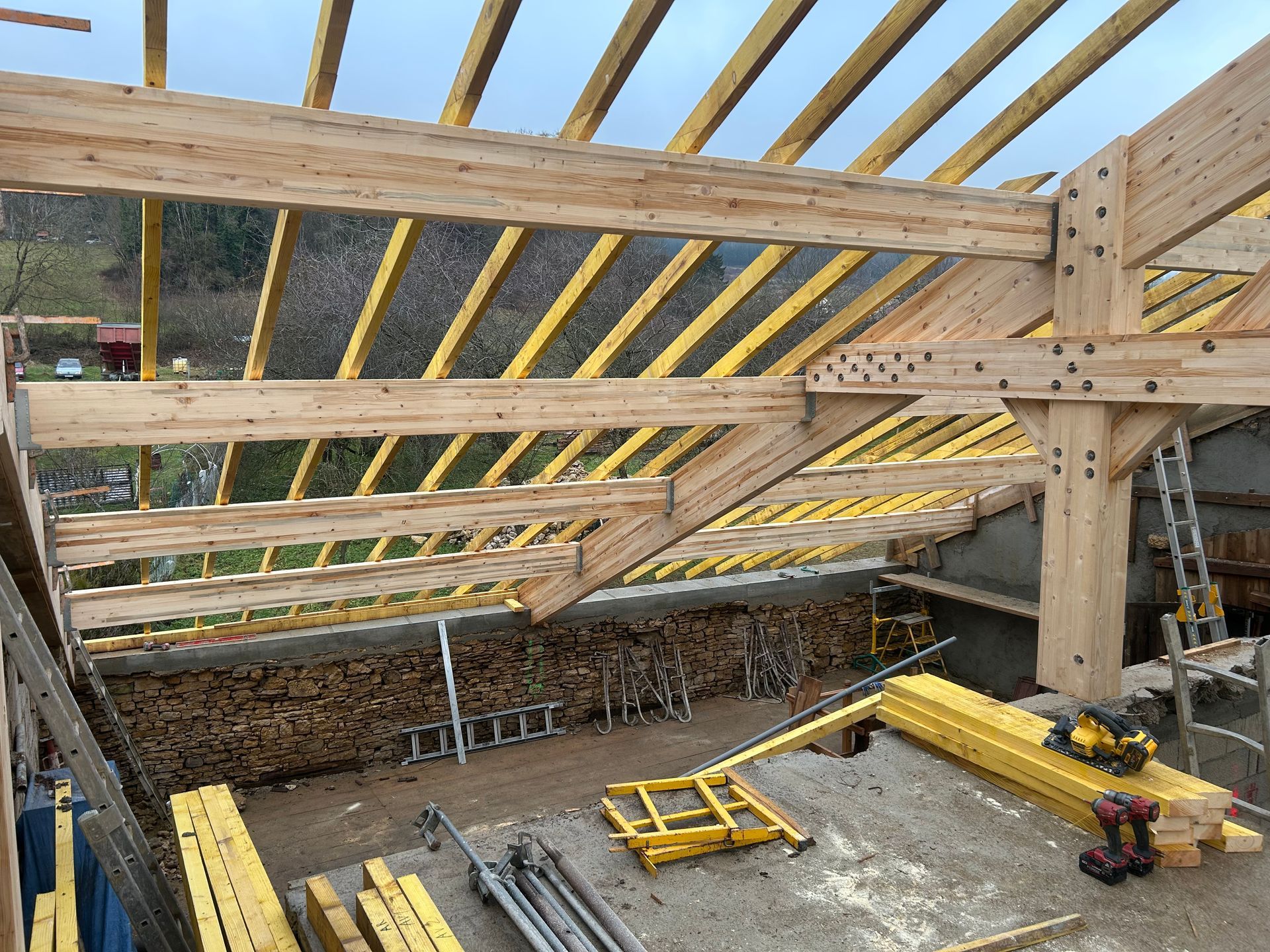 Une charpente de toit en bois inachevée, en construction, avec des chevrons, des poutres et du bois de chauffage empilé en dessous.