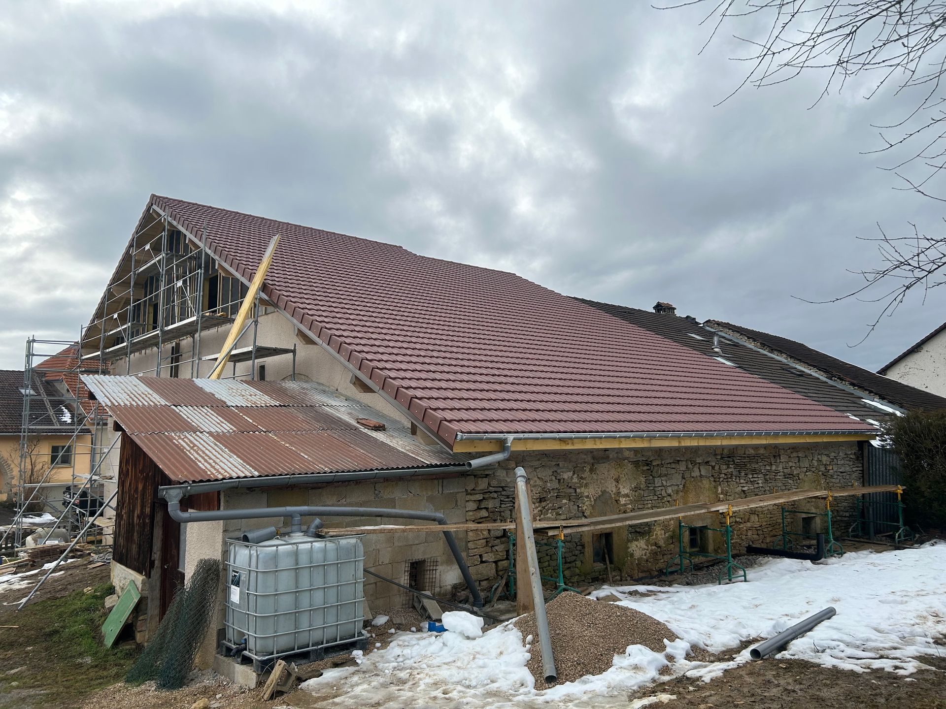 Une grange en pierre en cours de rénovation avec un toit de tuiles rouges fraîchement posé, une extension en métal rouillé et un réservoir d'eau sous la neige.