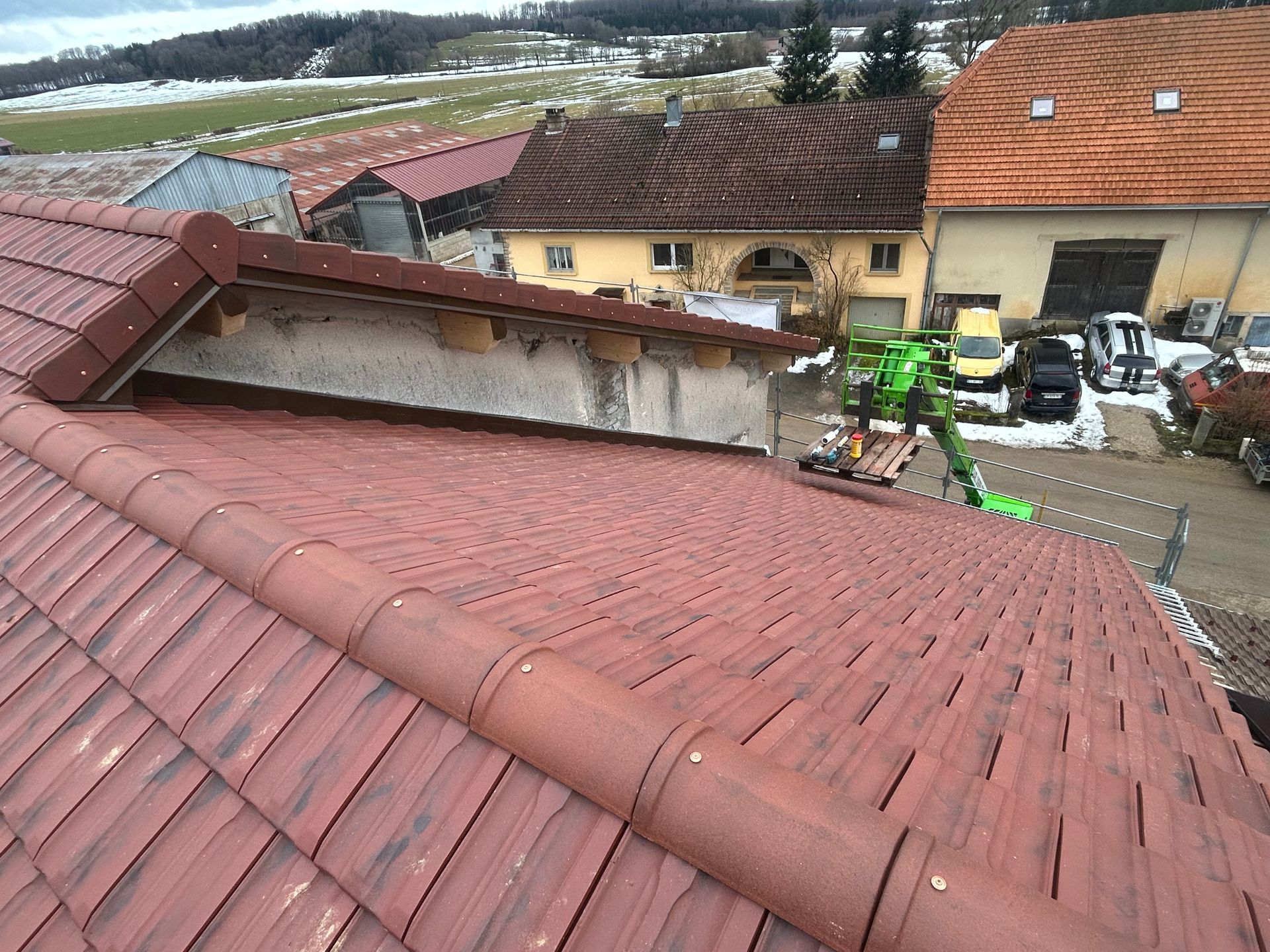 Vue en plongée d'un toit rouge fraîchement recouvert de tuiles, avec un espace inachevé et une petite grue verte en activité dans la cour en contrebas.