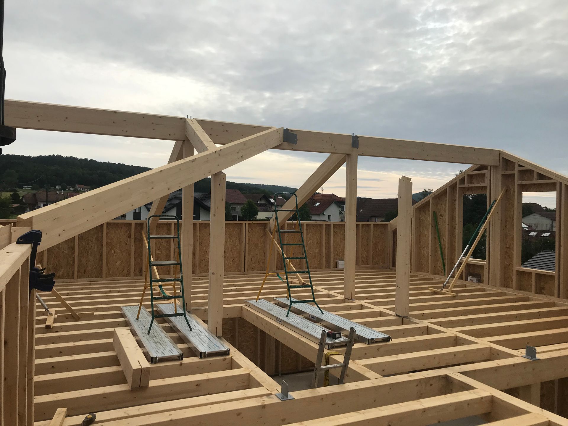 Chantier de construction montrant des ossatures murales en bois, des poutres de toit et des solives de plancher sous un ciel nuageux.