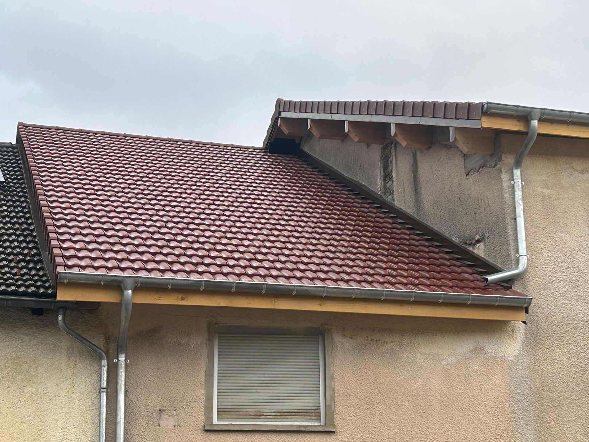 Gros plan sur l'extérieur d'une maison montrant un toit de tuiles rouges, un mur beige, une fenêtre avec un store fermé et des descentes de gouttière en métal.
