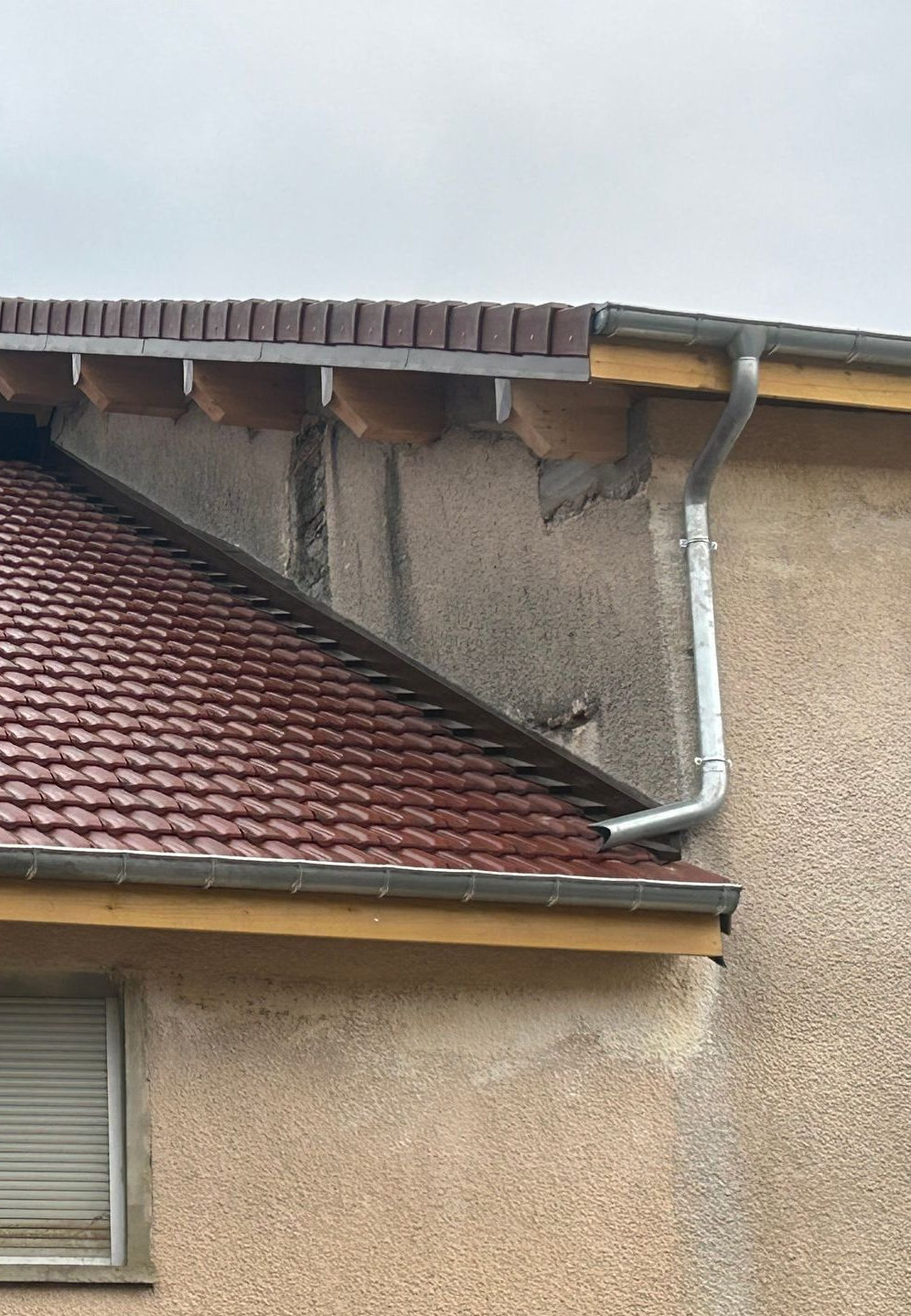 Vue latérale du toit d'une maison montrant une descente de gouttière en métal, des tuiles brunes et un mur en béton apparent avec une fissure verticale.