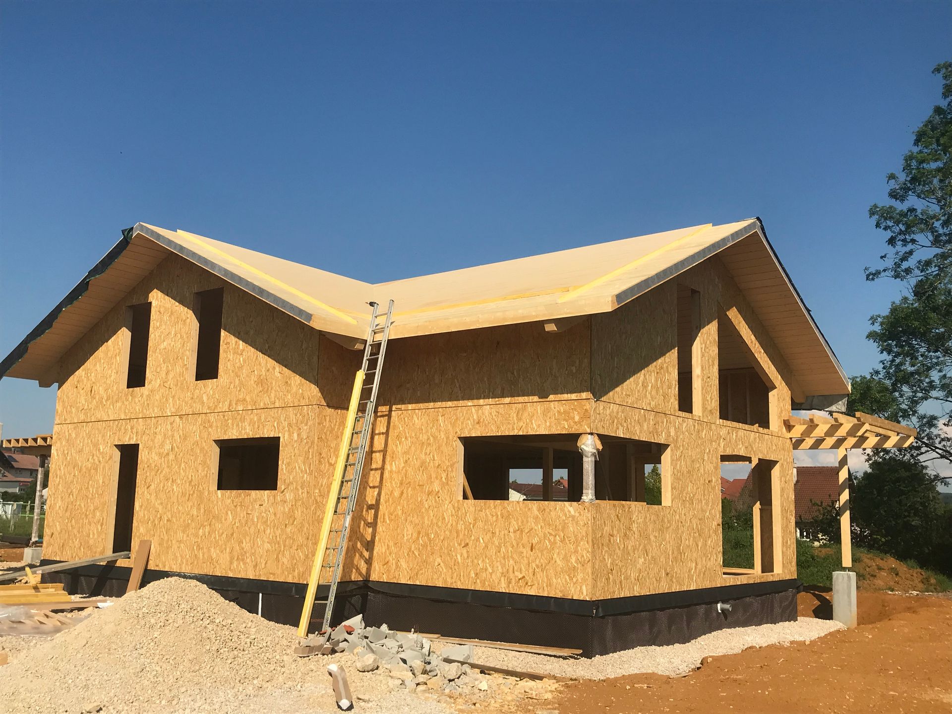 Une maison à deux étages en construction, avec des murs extérieurs en panneaux OSB, un toit en bois et une échelle appuyée contre le mur.