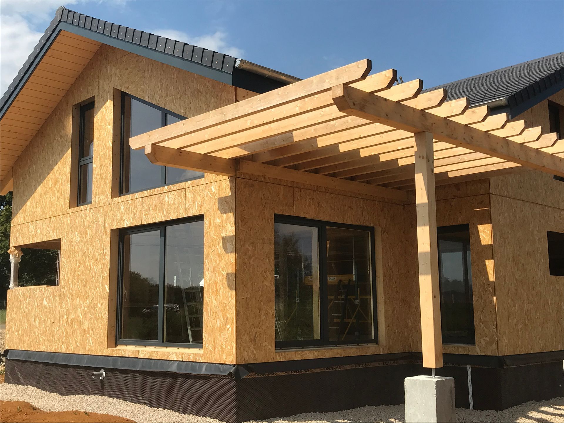 Une maison en construction avec des panneaux OSB en bois clair, de grandes fenêtres vitrées, un toit foncé et une pergola en bois à l'extérieur.