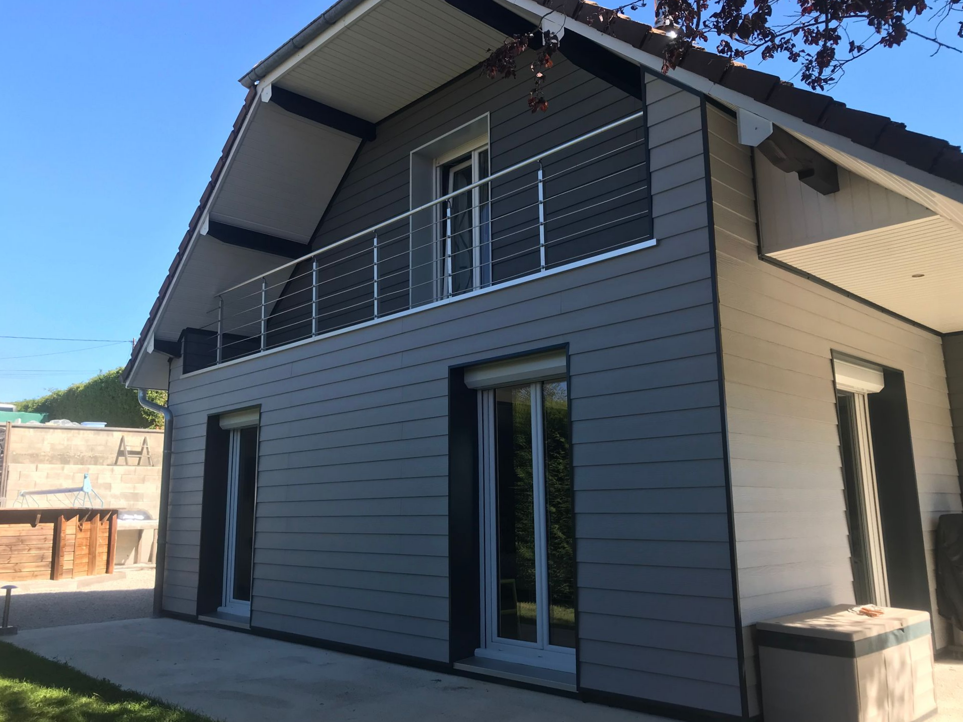 Une maison moderne de deux étages avec un bardage horizontal gris foncé, des boiseries blanches, un balcon en métal et de grandes portes-fenêtres donnant sur une terrasse.