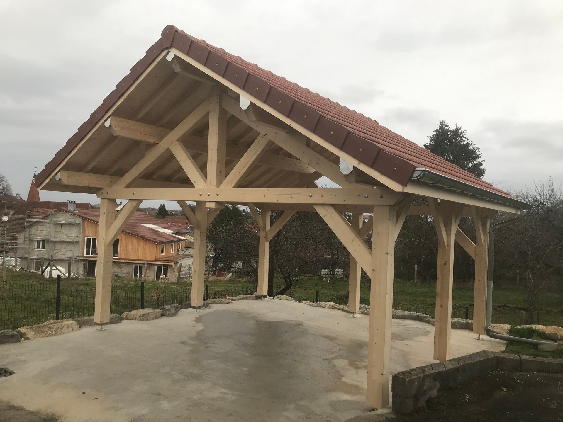Une structure à ossature en bois ouverte sur les côtés, coiffée d'un toit à pignon en tuiles rouges, repose sur une dalle de béton dans une cour rurale.