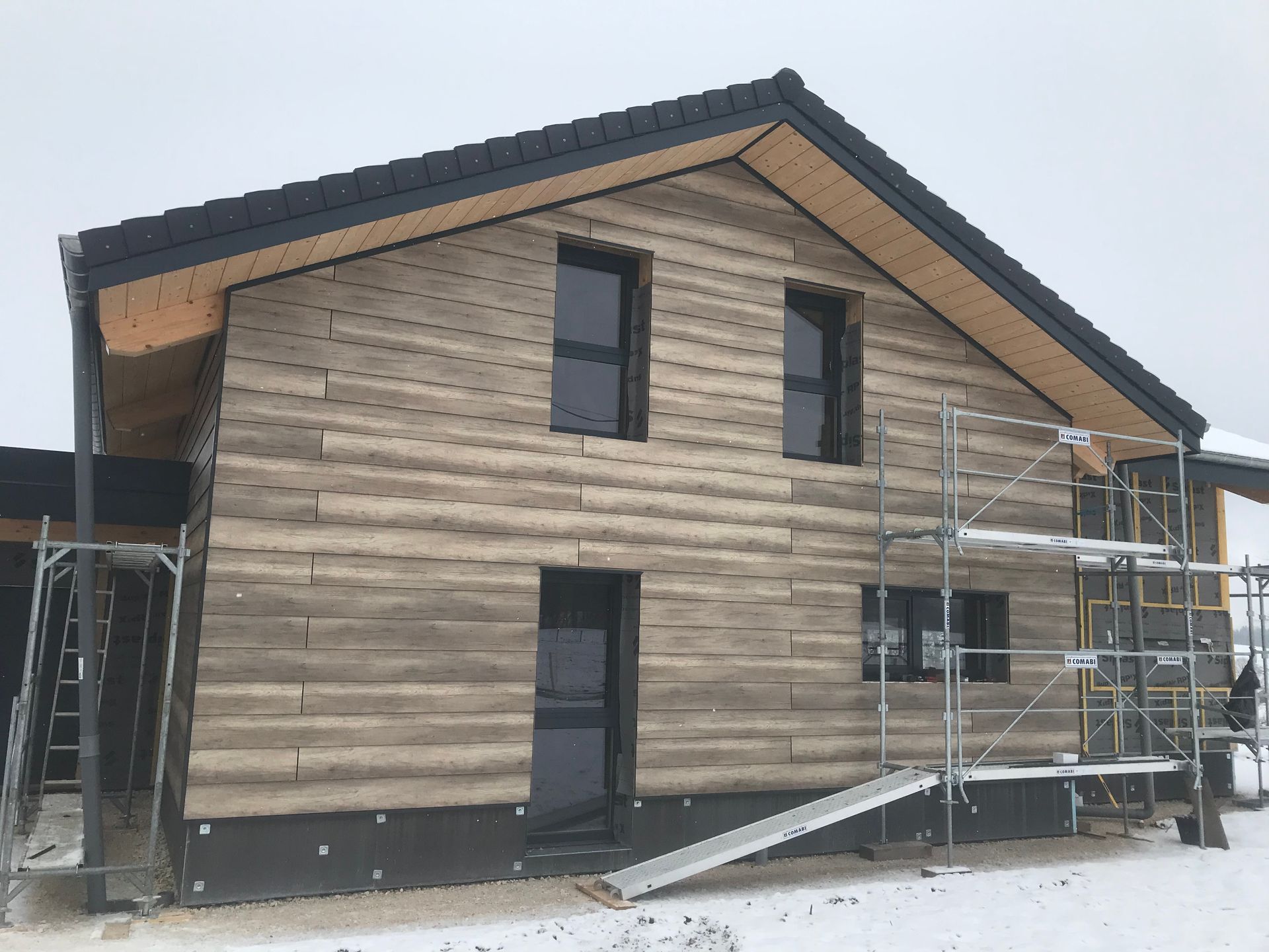 Une maison à deux étages en construction, avec un bardage beige imitant le bois, un toit sombre et des échafaudages métalliques, par une journée enneigée.