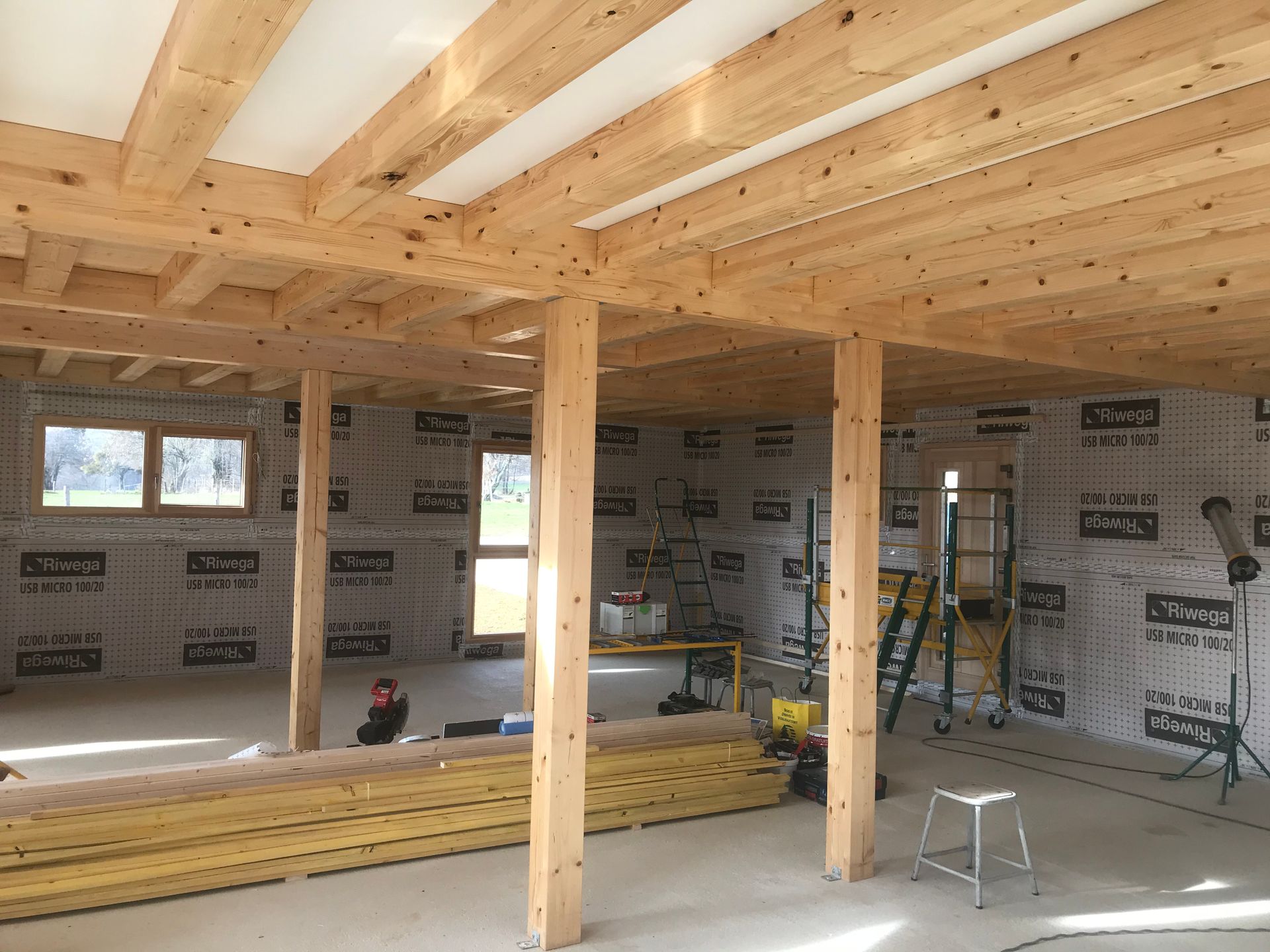 Intérieur d'un bâtiment en construction présentant des poutres en bois apparentes, des poteaux de soutien et des murs recouverts d'un pare-vapeur.
