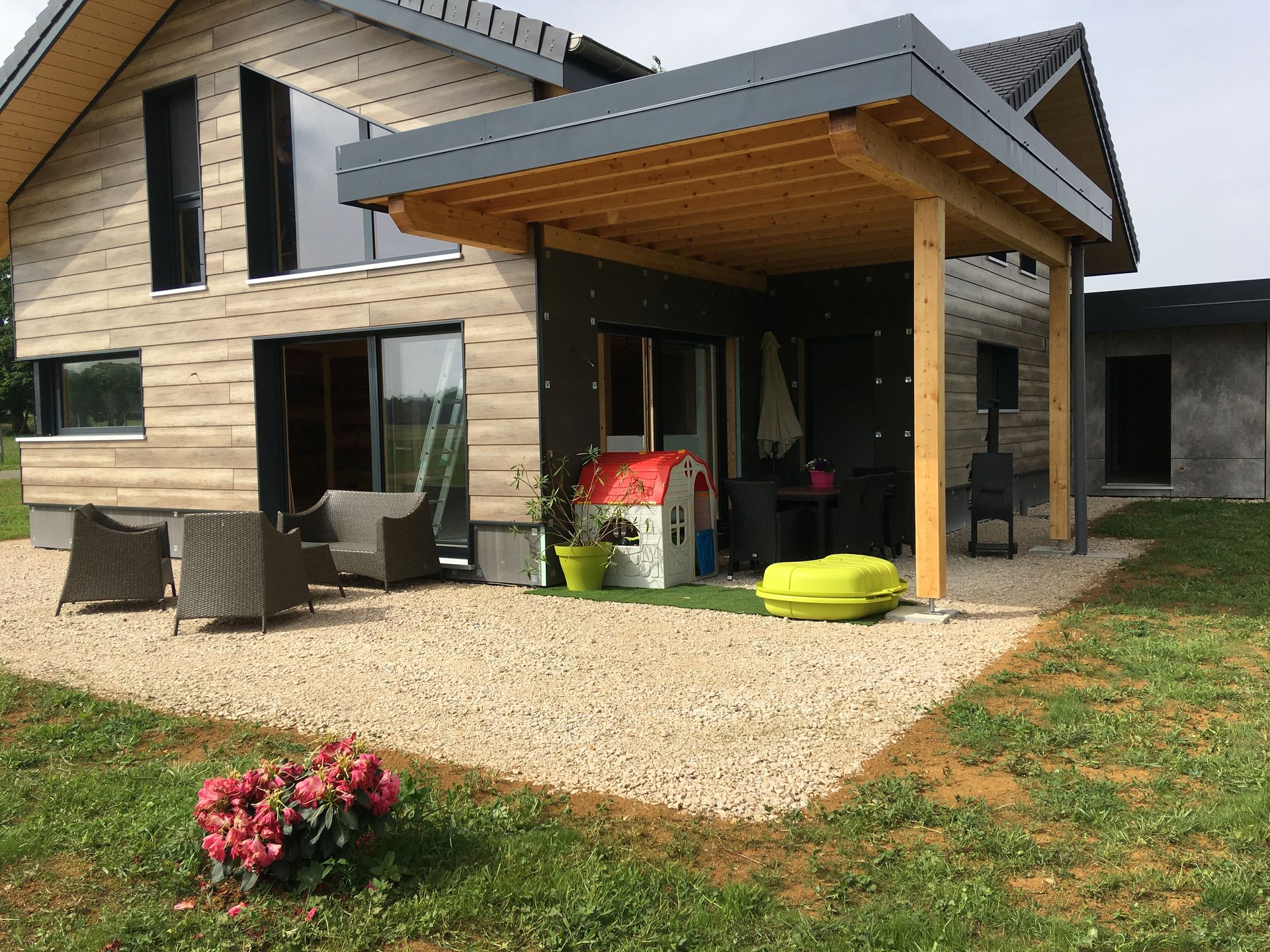Une maison moderne à deux étages avec des lambris clairs, une grande terrasse en gravier et une pergola en bois couverte.