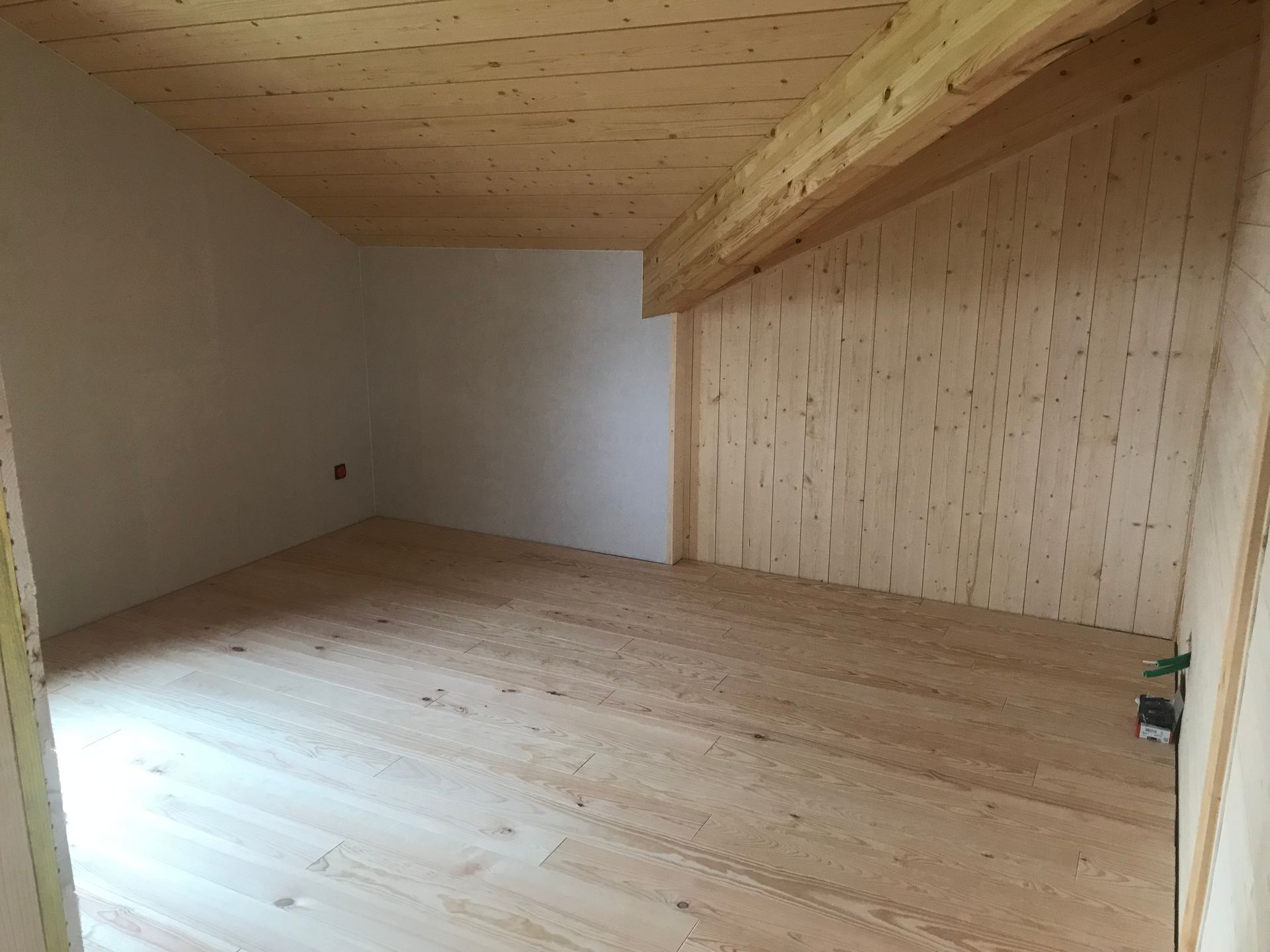 Une pièce vide avec un parquet clair, un plafond en bois incliné et des murs mêlant plâtre blanc et lambris.