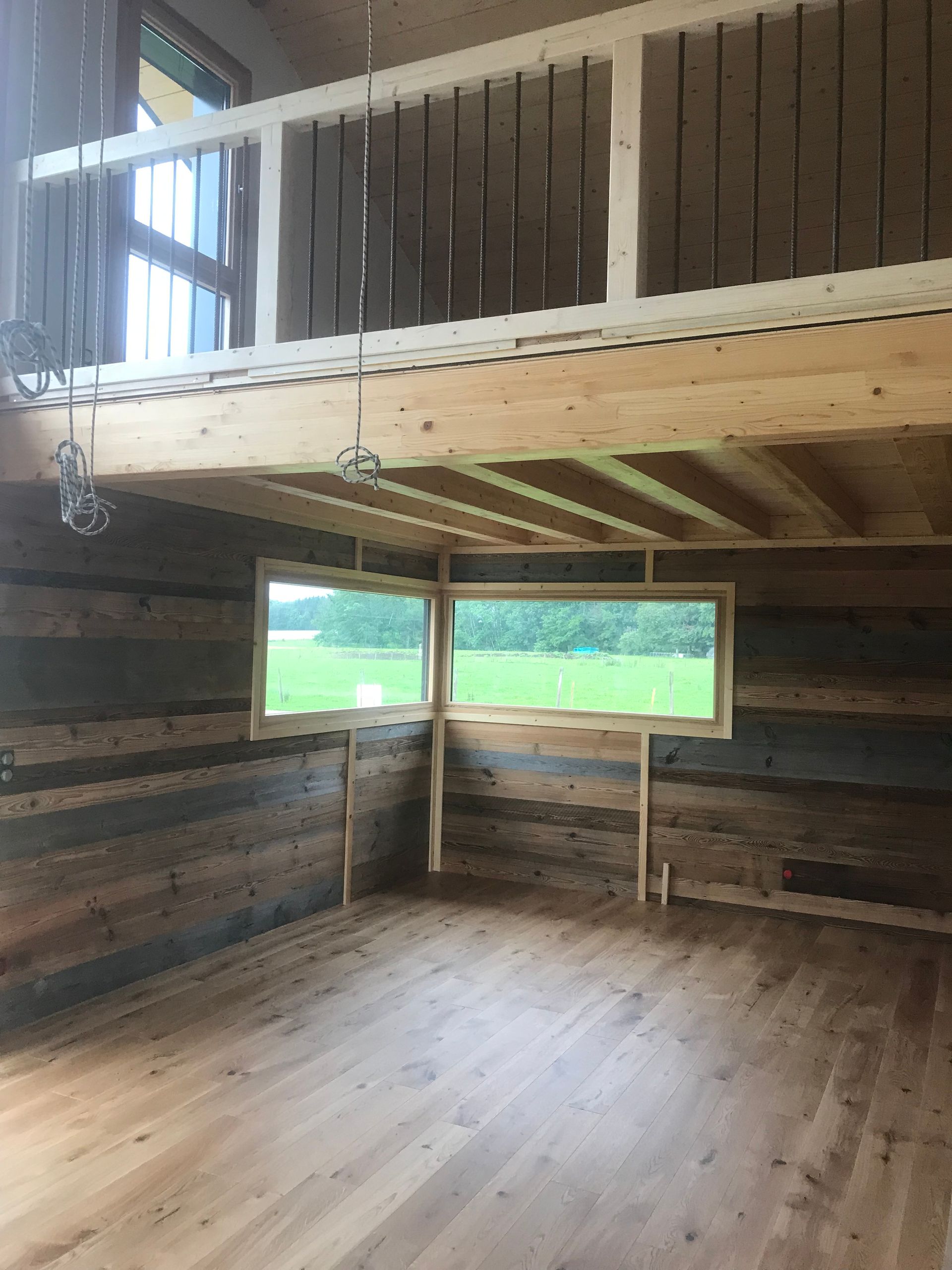 Une pièce rustique avec des murs en rondins horizontaux, un plancher en bois et une mezzanine avec une balustrade blanche donnant sur un champ verdoyant.