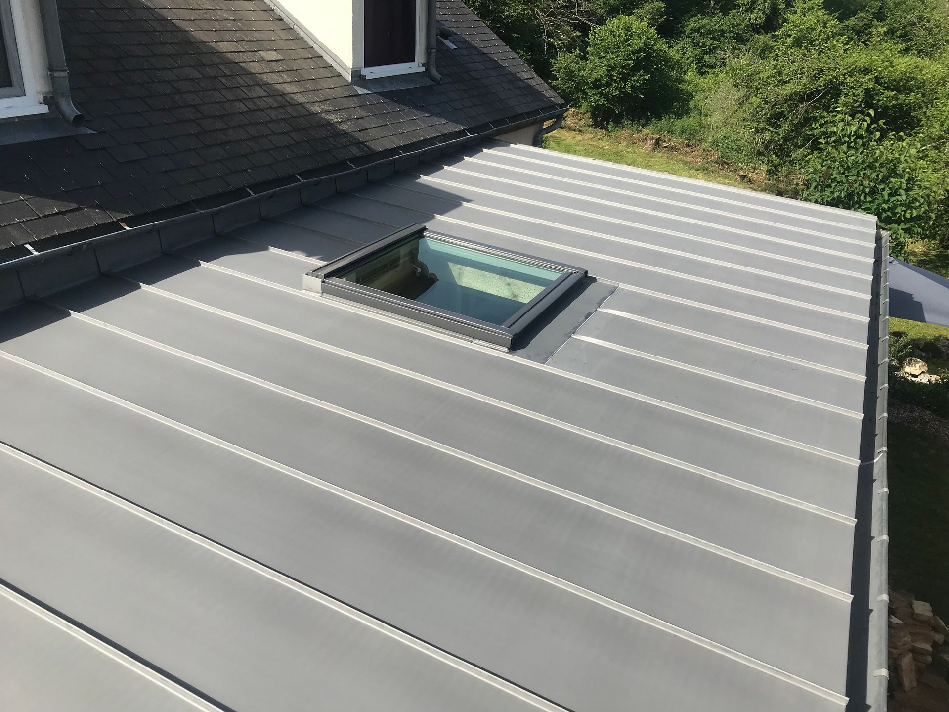 Une extension de toiture en métal gris à joints debout, avec un puits de lumière rectangulaire installé, est fixée à une maison aux tuiles foncées.