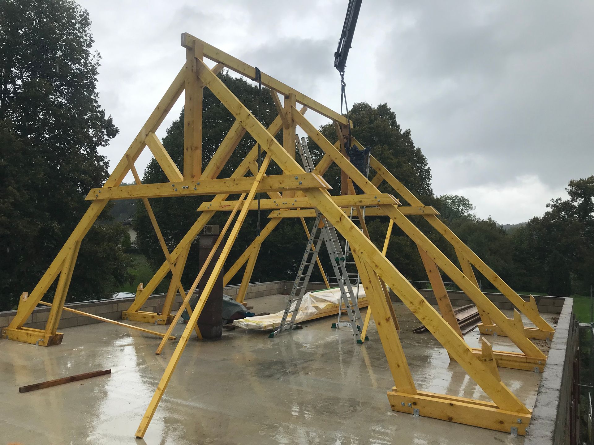 Une grue soulève une charpente de toit en bois et la pose sur des fondations en béton à l'extérieur, sous un ciel nuageux.