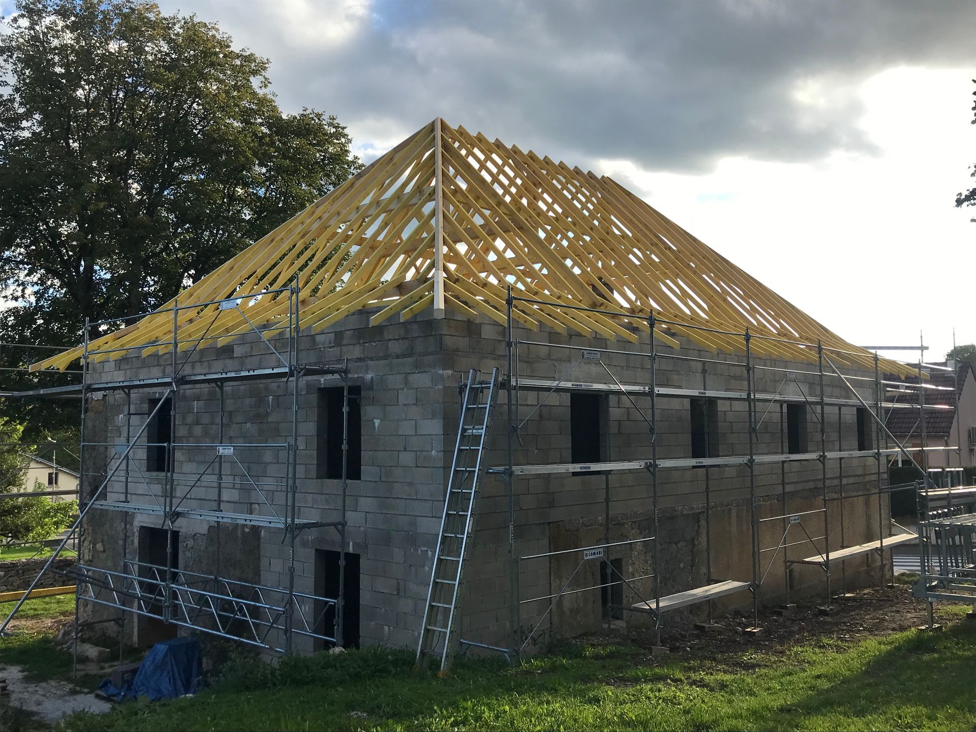 Un bâtiment en pierre de deux étages en construction, avec une charpente de toit en bois jaune apparente et des échafaudages tout autour.
