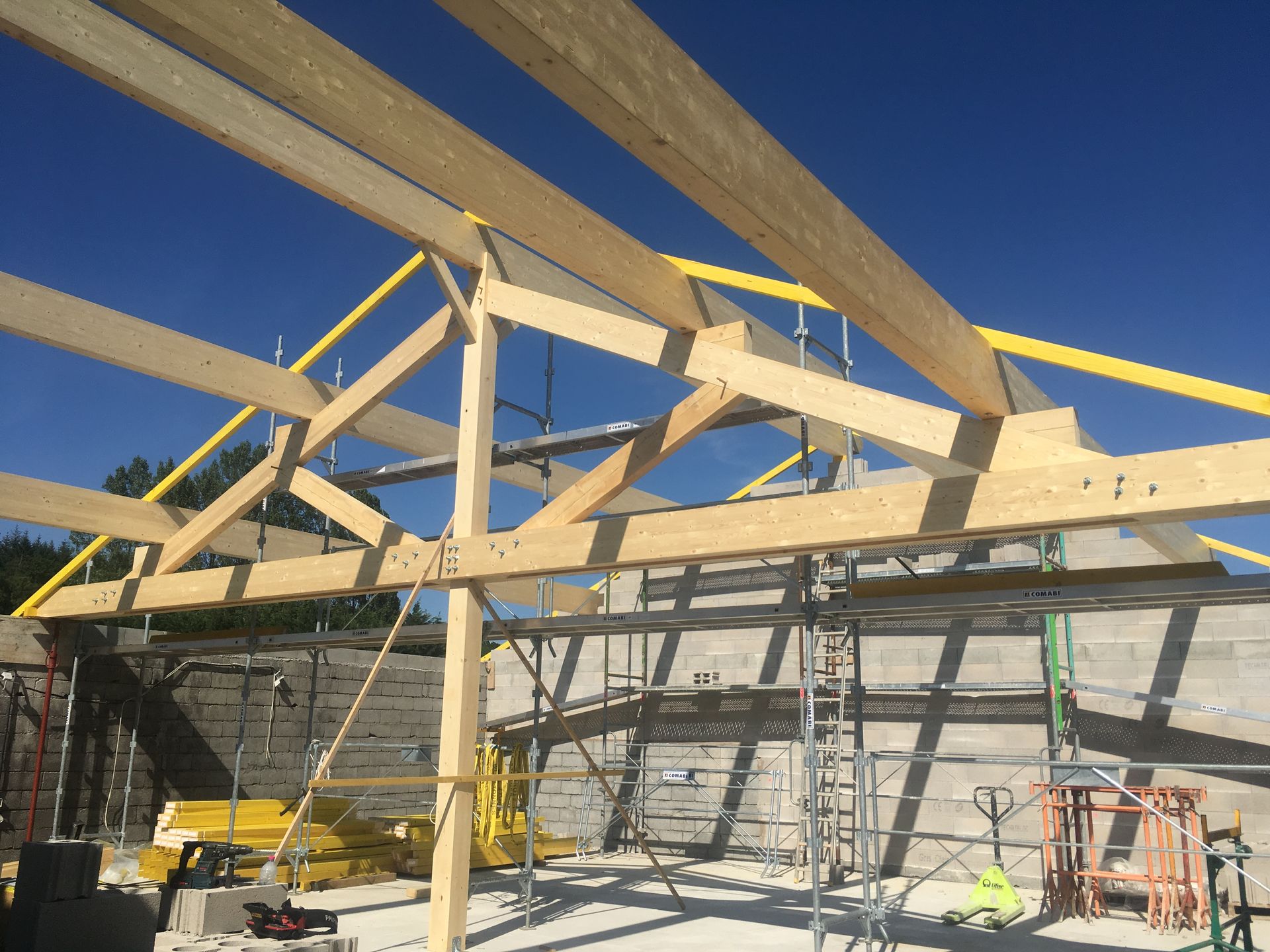 Une charpente de toit en bois en construction se détache sur un ciel bleu clair, avec des échafaudages visibles en arrière-plan.