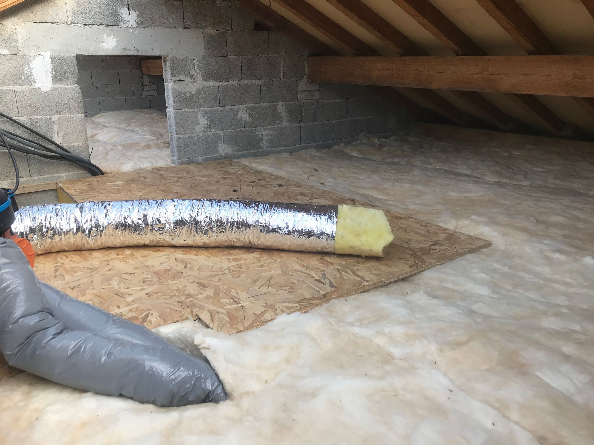 Combles isolés avec murs en parpaings apparents, chevrons en bois et conduit de ventilation CVC sur une base en bois.