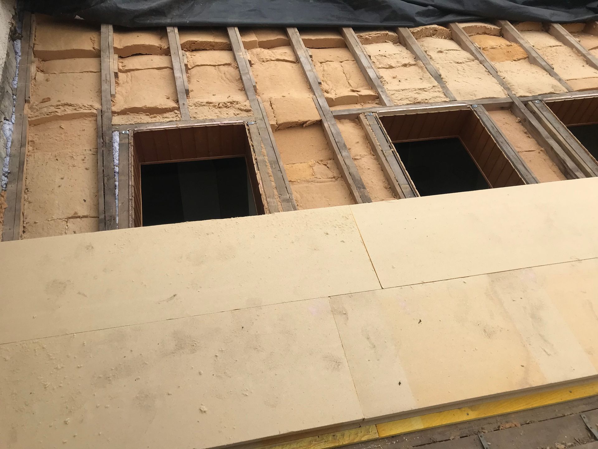 Des panneaux isolants sont installés entre les chevrons du toit, laissant apparaître des espaces vides, et un nouveau panneau est en cours de pose au premier plan.