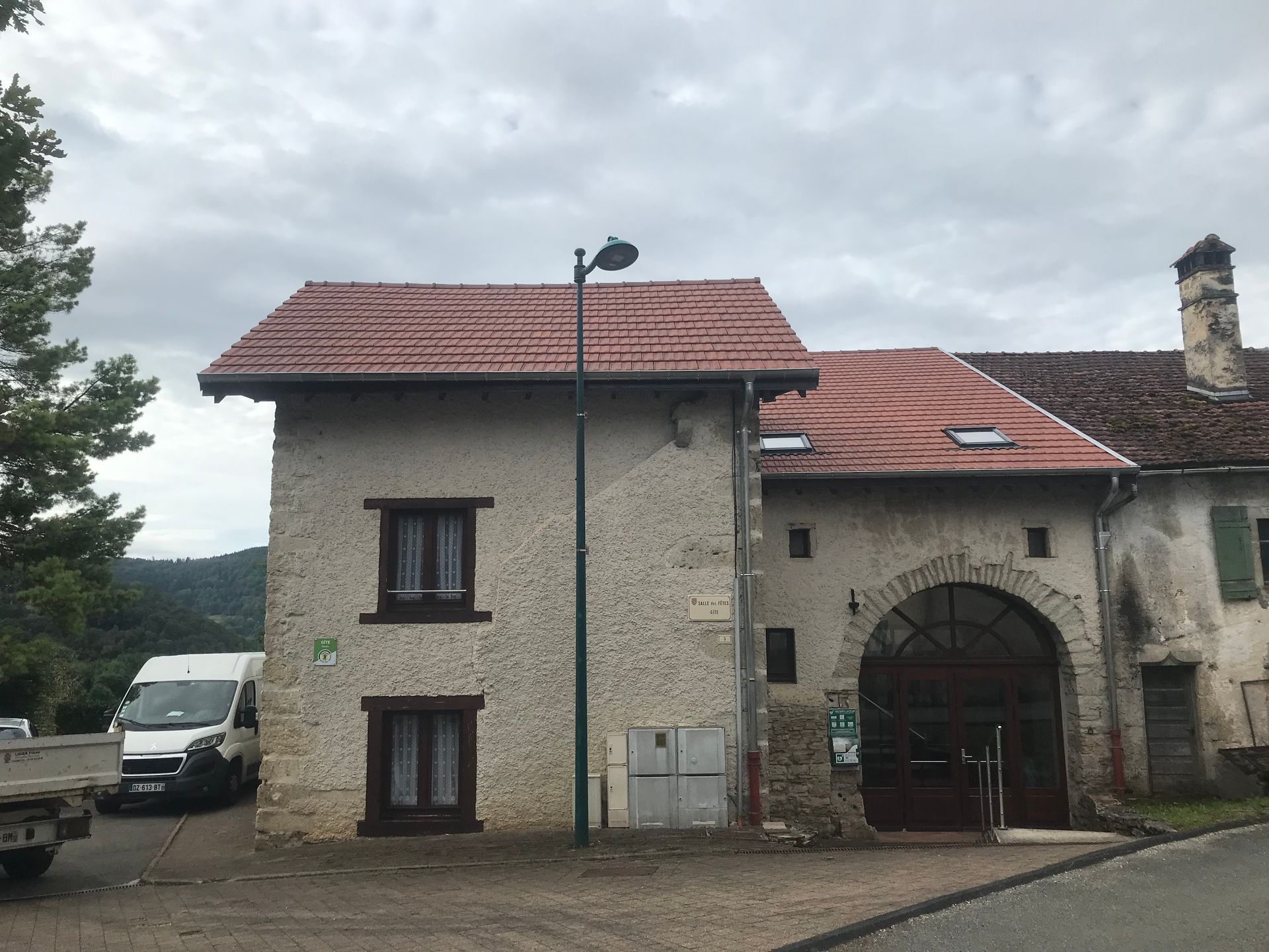 Un bâtiment rustique en pierre, avec un toit de tuiles rouges et une entrée voûtée, se dresse à côté d'un lampadaire par temps nuageux.