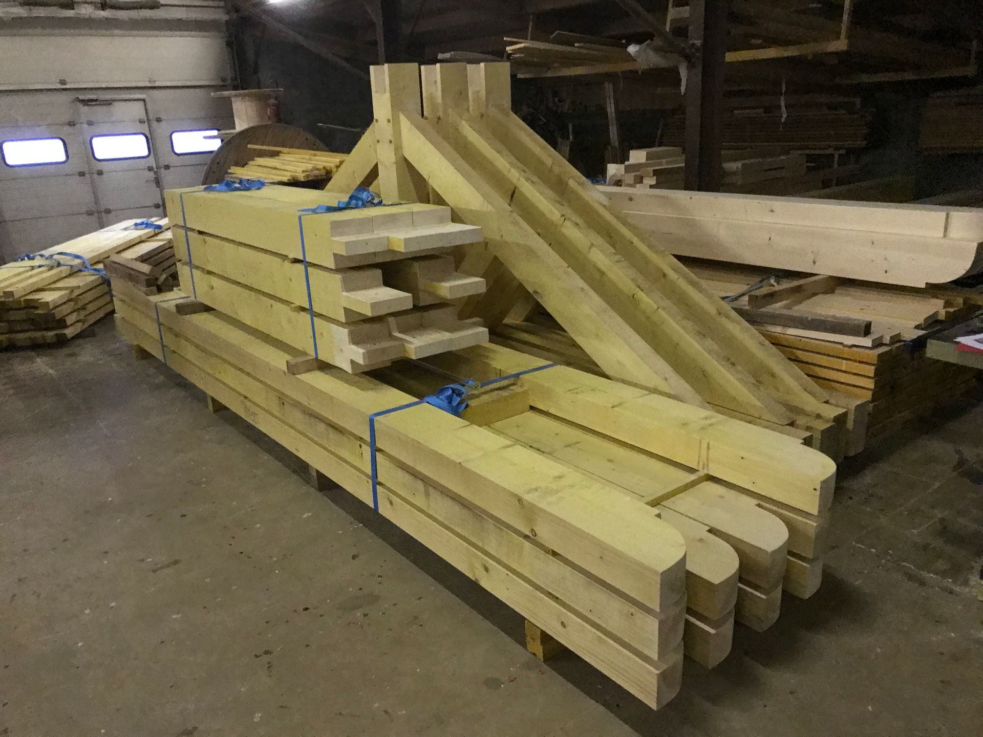 Des poutres en bois clair empilées et des éléments de charpente sur mesure sont stockés dans un atelier.