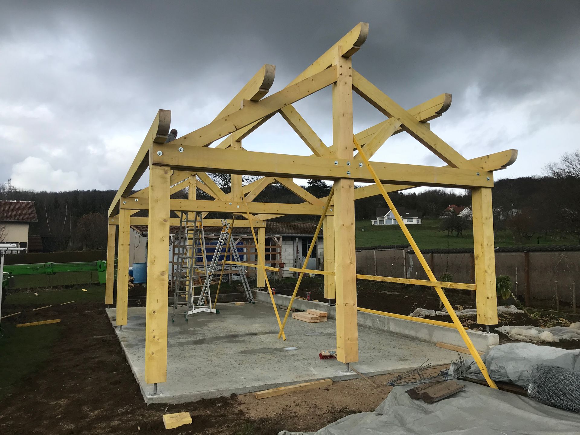 Un pavillon à ossature en bois partiellement construit, avec des poutres en bois jaunes, reposant sur des fondations en béton, dans une zone rurale.
