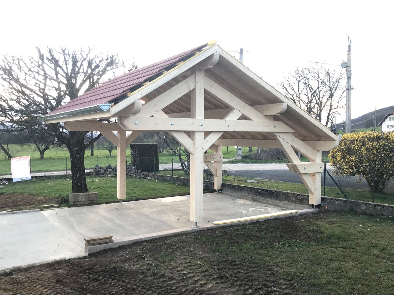 Un abri de voiture en bois nouvellement construit, avec un toit de tuiles rouges, se dresse au-dessus d'une base en béton, dans un espace extérieur herbeux et dégagé.