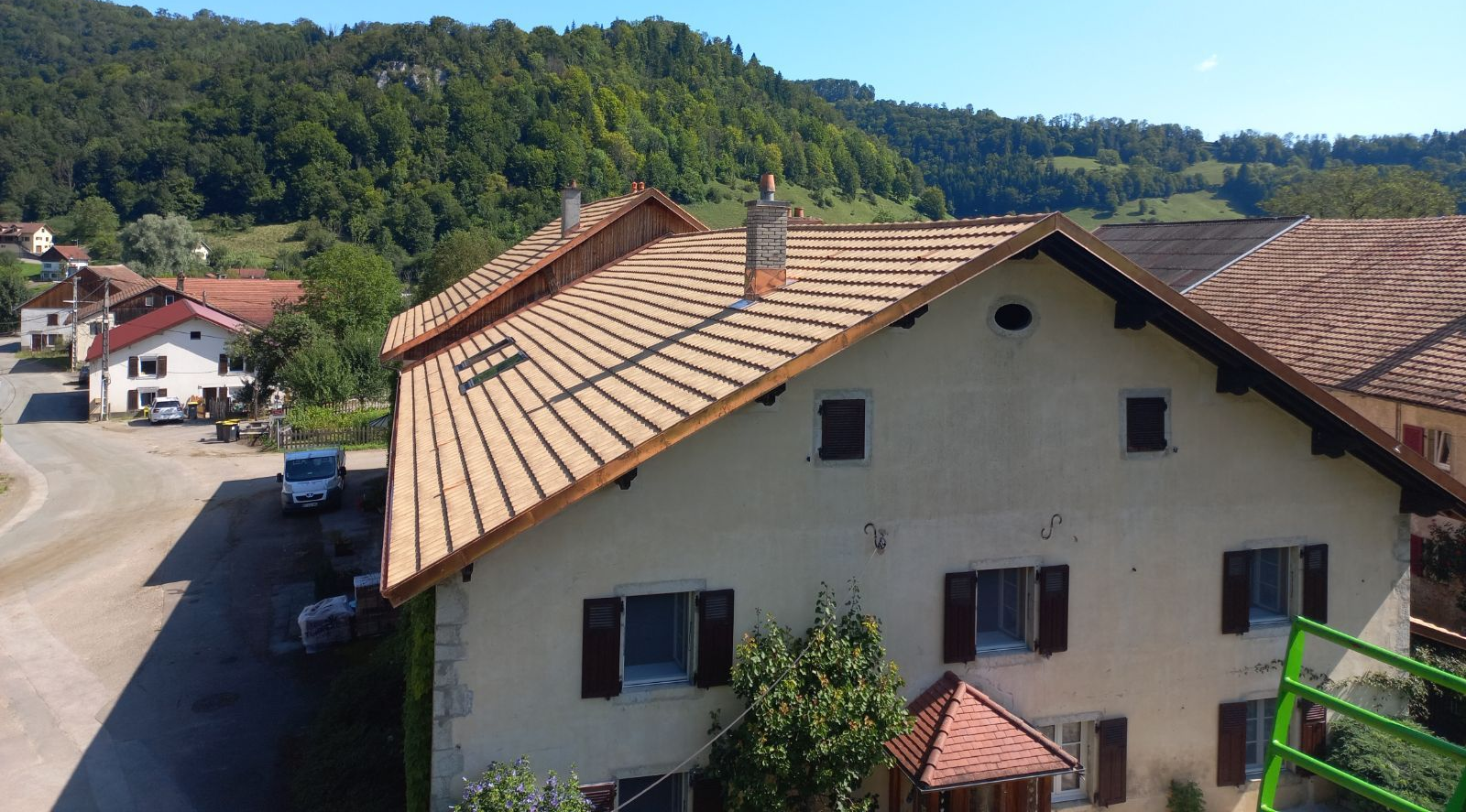 Une vue aérienne montre une maison de couleur claire avec un toit neuf en tuiles brun clair, au bord d'une route dans un paysage vallonné et verdoyant.