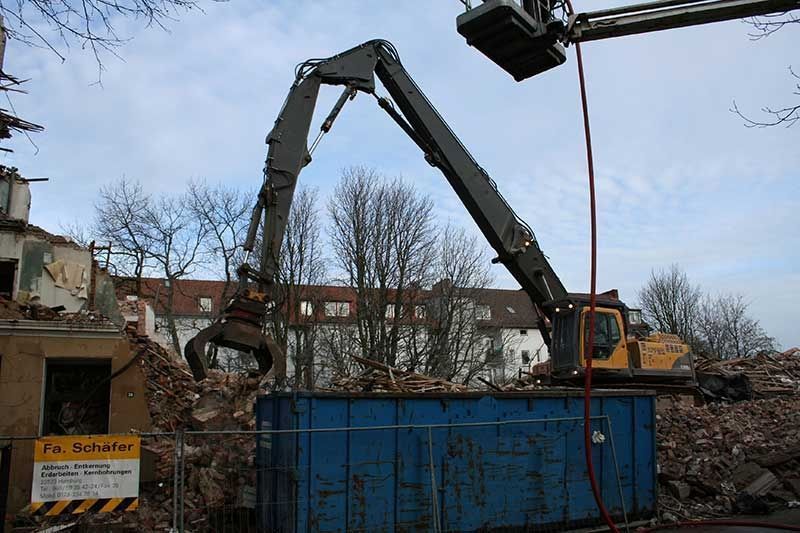Ein Abbruchbagger lädt Schutt in einen blauen Container auf einer Baustelle.