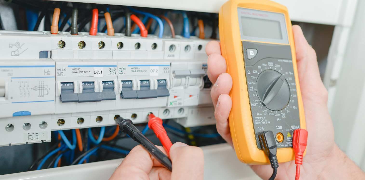 Una persona sostiene un multímetro delante de una caja eléctrica.