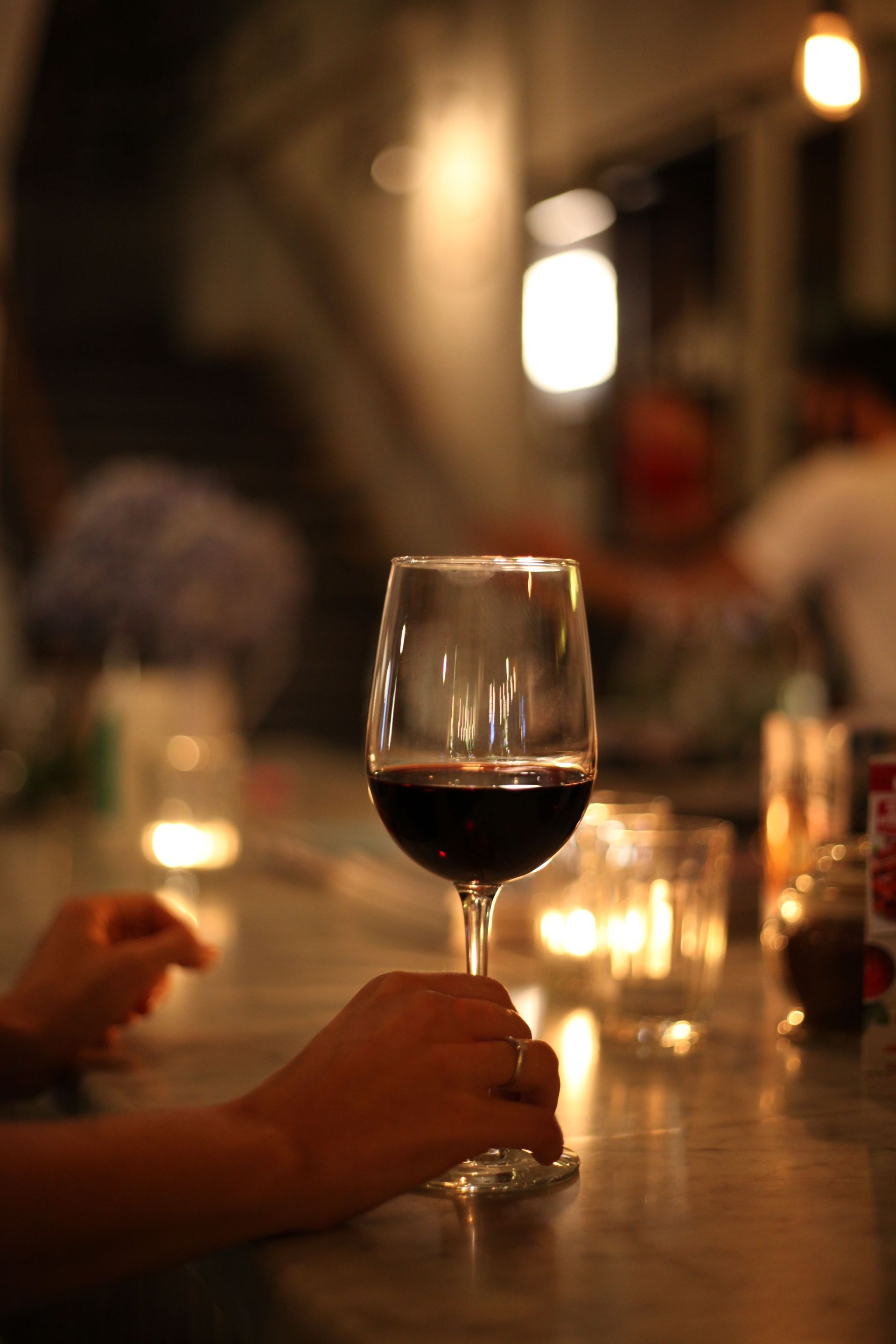 Una copa de vino sobre una mesa de restaurante con poca luz, con velas y comensales borrosos al fondo.