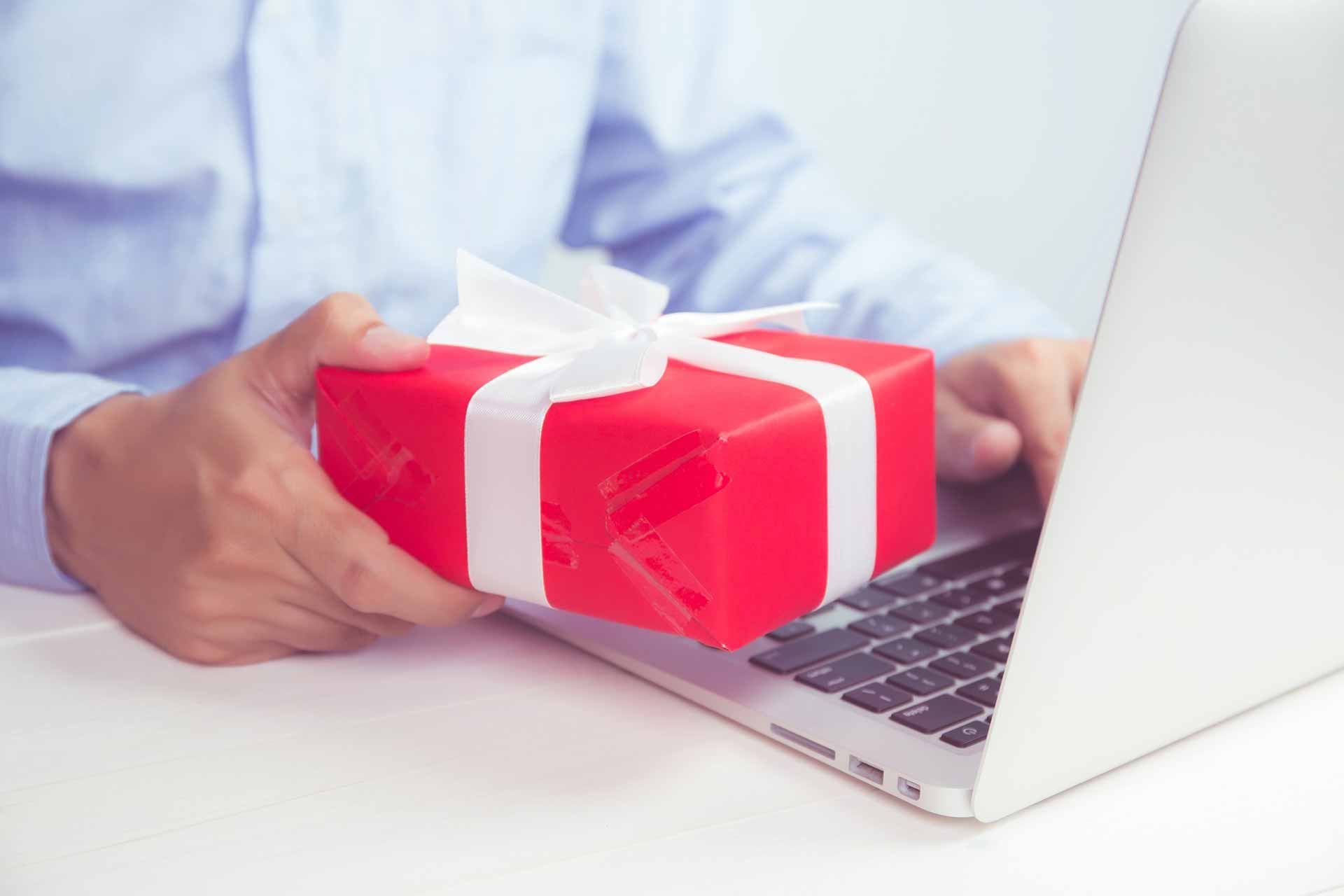 Un hombre sostiene una caja de regalo roja frente a una computadora portátil.