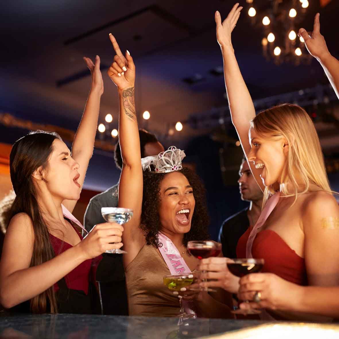 Un grupo de mujeres está sentada en un bar sosteniendo copas de vino y levantando los brazos en el aire.
