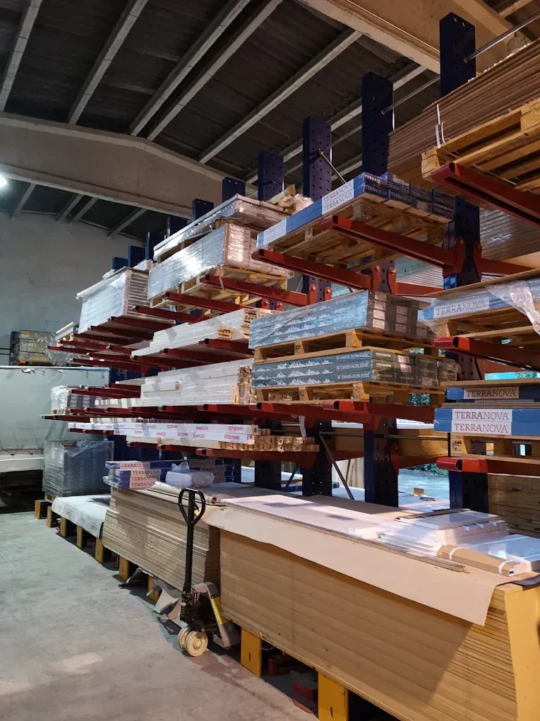 Estanterías de almacén apiladas con largas tablas de madera y materiales sobre palés en un interior industrial.