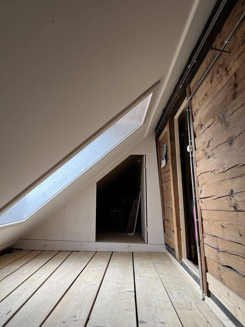 Dachboden mit schräger Decke, freiliegenden Holzwänden und unfertigen Dielen, die zu einer dunklen Tür führen