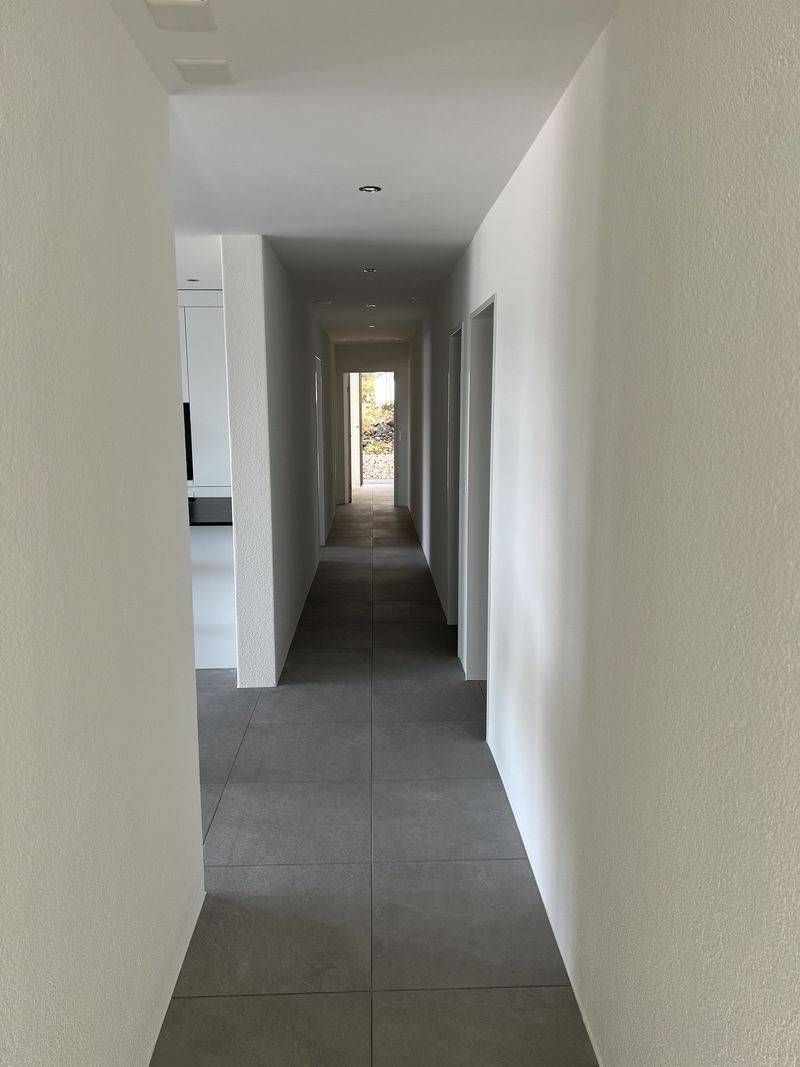 Ein schmaler, weisser Flur mit grauem Teppich führt zu einer hellen Tür am anderen Ende.