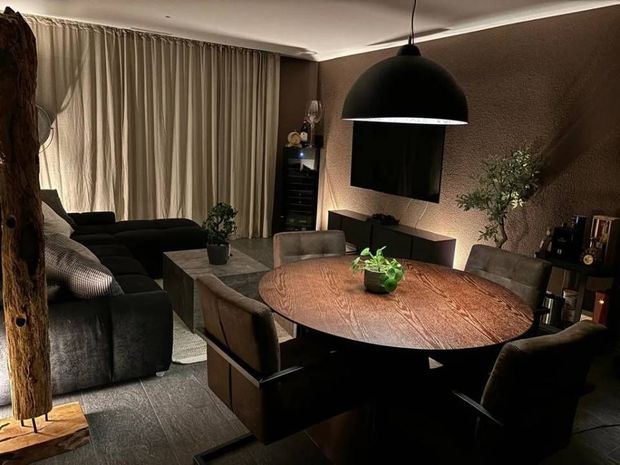 Gemütliches Wohnzimmer mit dunklem Sofa, rundem Esstisch, Lampe, Vorhängen und warmen braunen Wänden
