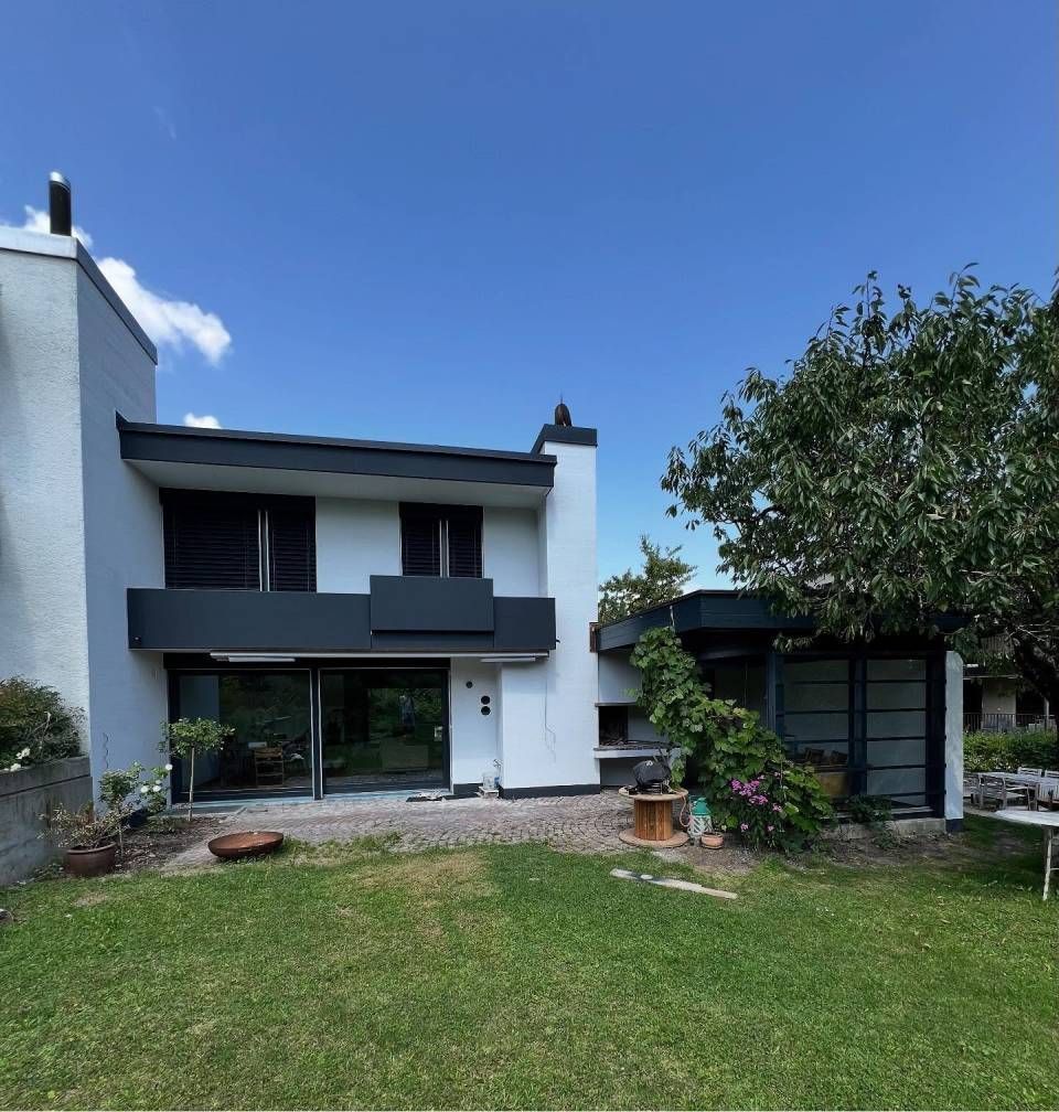 Zweistöckiges modernes Haus mit weissen Wänden, dunklem Balkon und grünem Rasen unter blauem Himmel