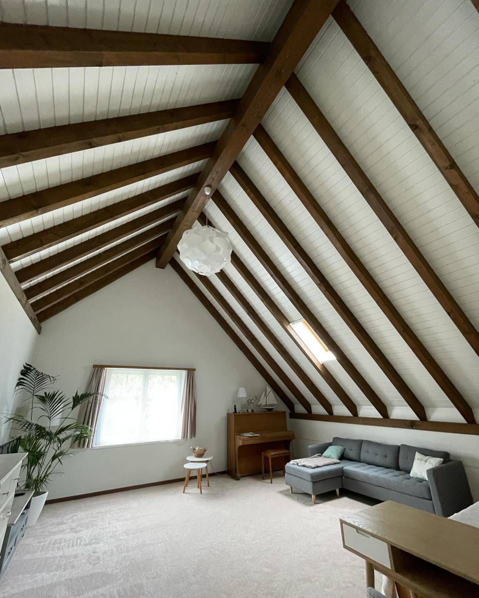 Ein helles Dachgeschosswohnzimmer mit freiliegenden Holzbalken, grauem Ecksofa, Fenster, Klavier und weisser Deckenleuchte.