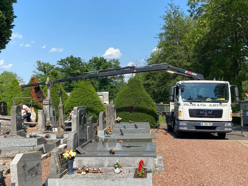 Un ouvrier, à bord d'un camion utilitaire blanc, utilise une grue déployée parmi les pierres tombales d'un cimetière.