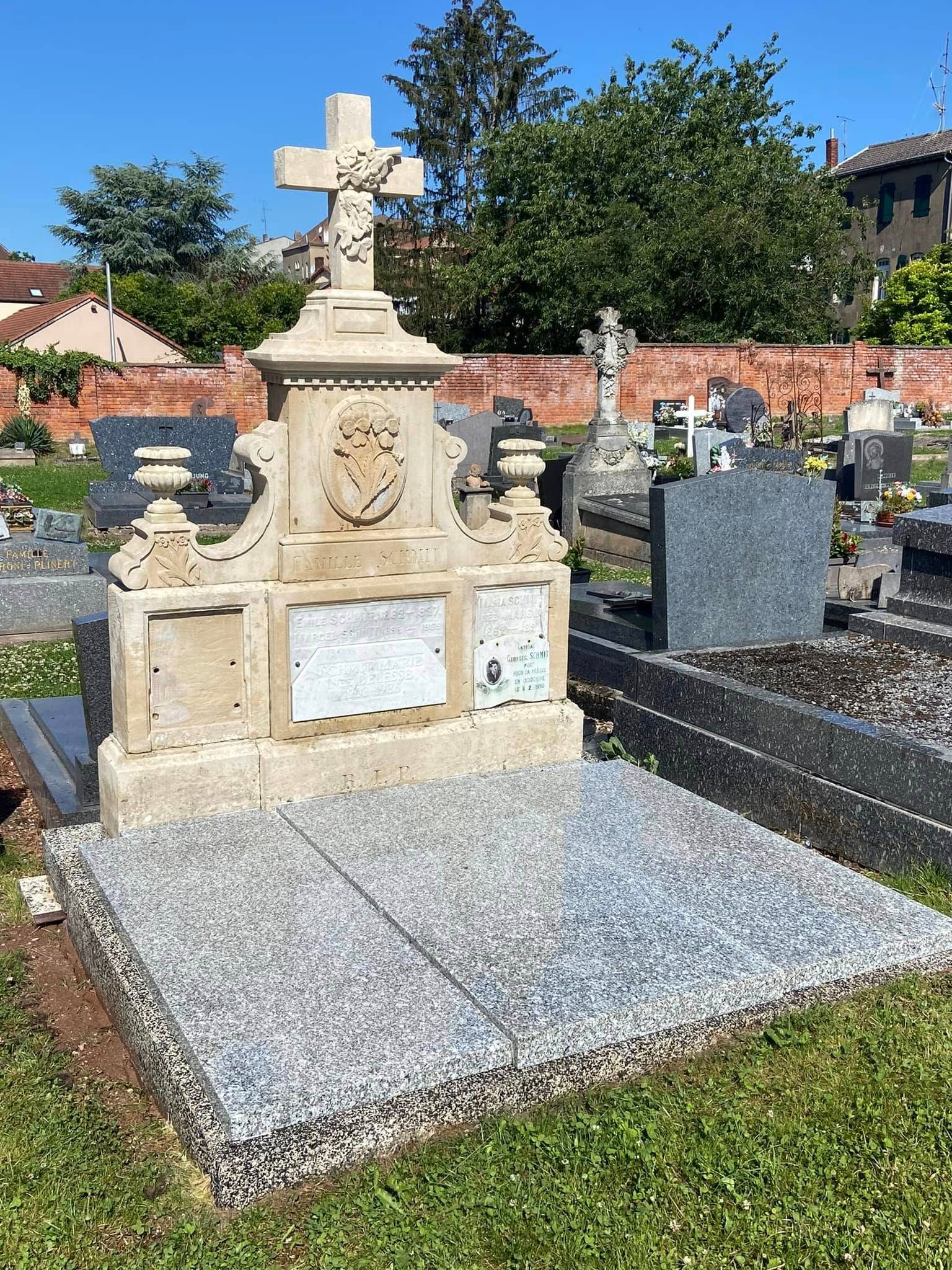 Pierre tombale en pierre ornée d’une croix dans un cimetière, entourée d’autres pierres tombales et d’un mur de briques.