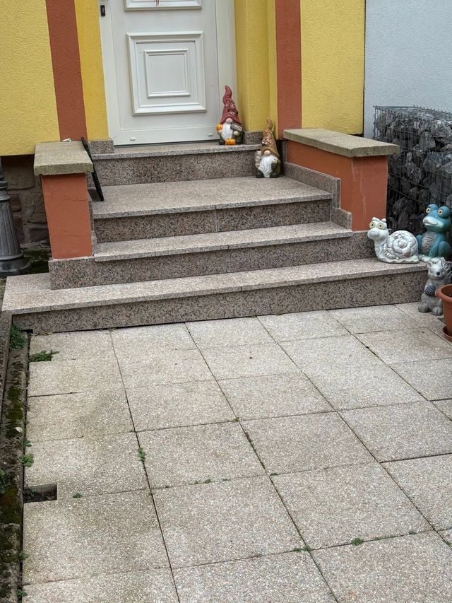 Un perron et une cour carrelée mènent à une porte blanche, ornée de petits pots de fleurs près de l'entrée.