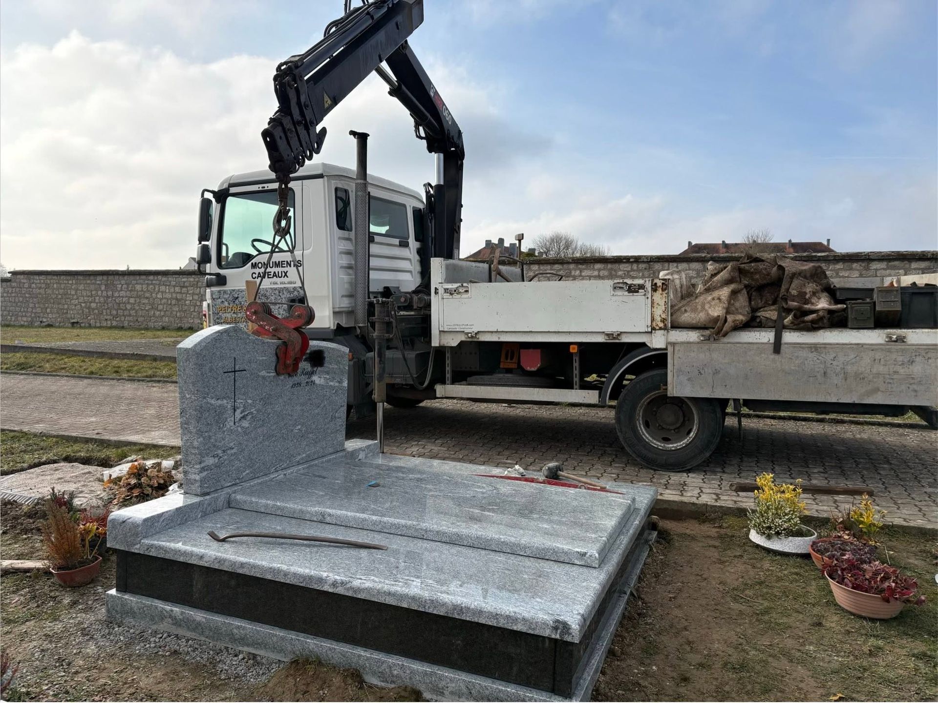 Camion-grue soulevant une stèle funéraire en pierre grise dans un cimetière sous un ciel nuageux.