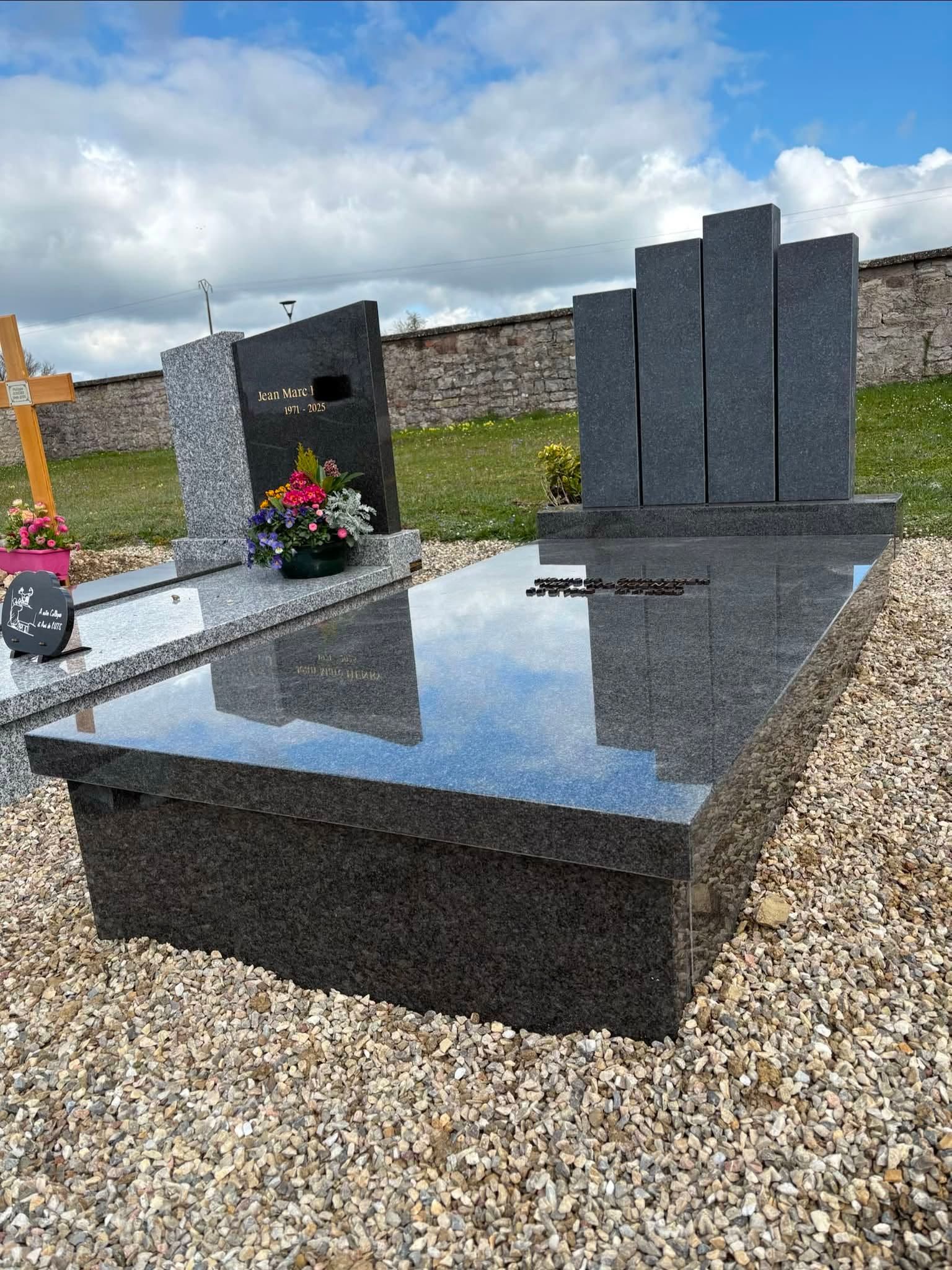 Pierre tombale en granit noir poli ornée de fleurs commémoratives dans un cimetière sous un ciel nuageux.
