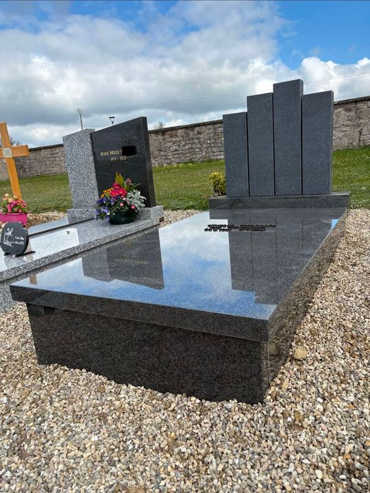Pierre tombale en granit noir à surface polie dans un cimetière, avec des fleurs et des pierres tombales à côté.