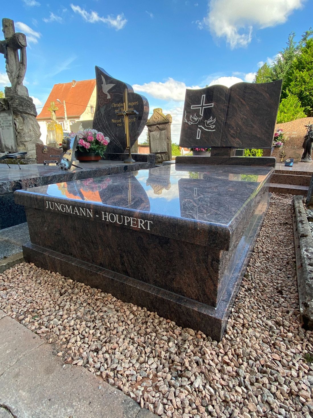 Pierre tombale en granit noir ornée de fleurs et d'une statue en pierre dans un jardin de cimetière ensoleillé.