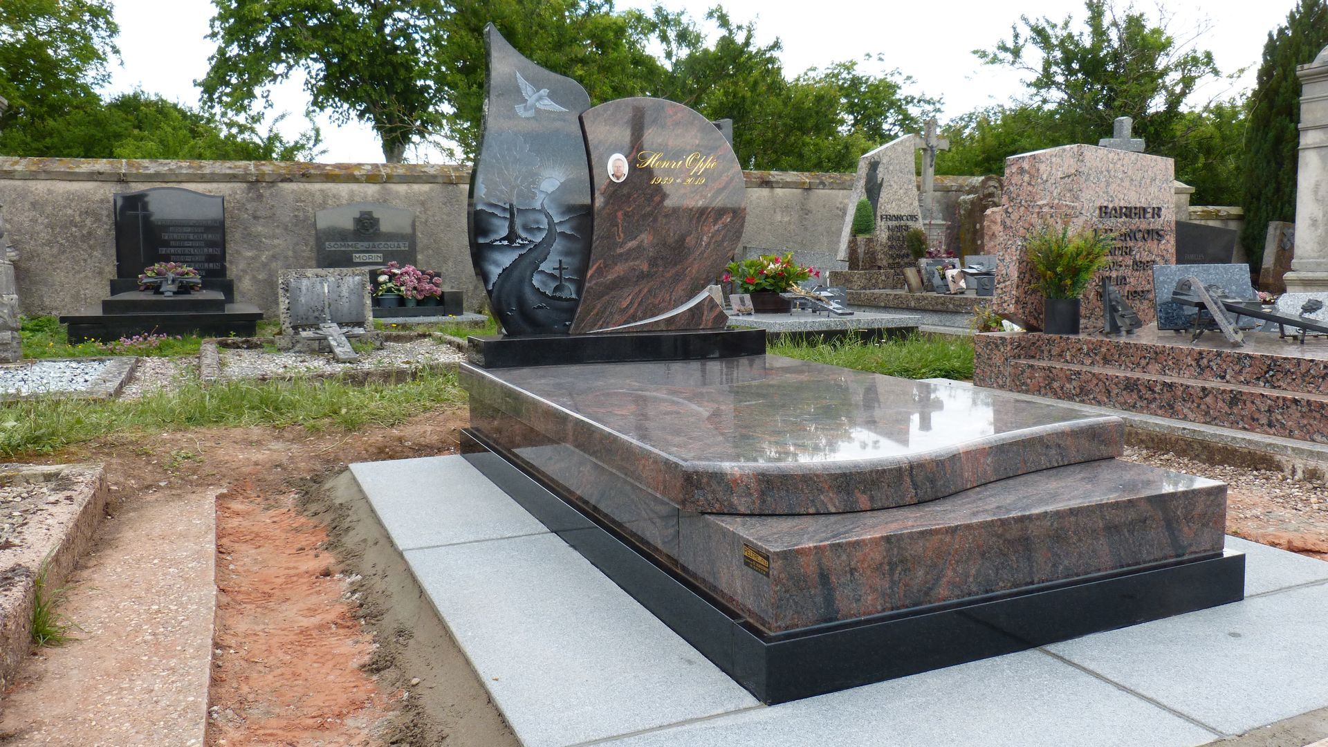 Cimetière avec pierre tombale en granit noir poli et sépulture entourée d'arbres et de pierres.