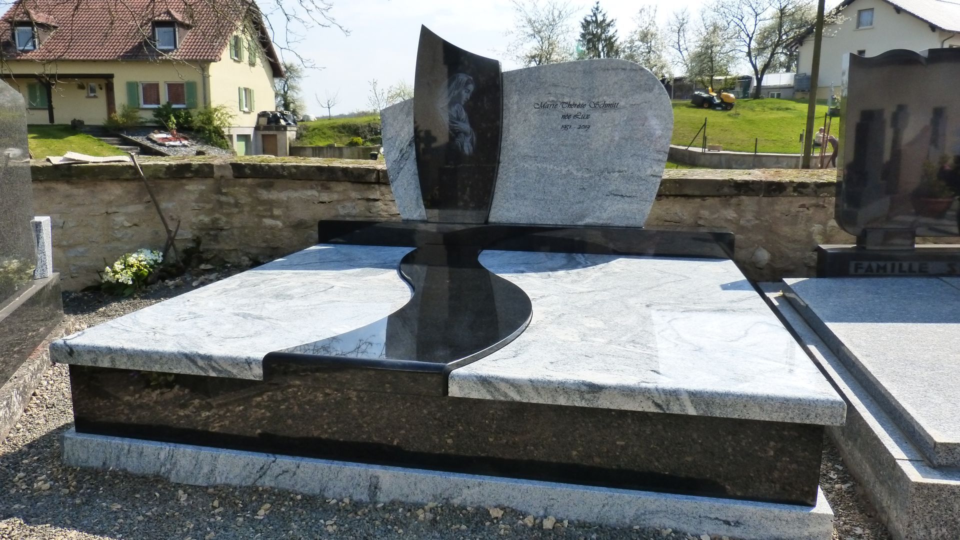 Monument funéraire moderne en granit noir et blanc dans un cimetière avec des maisons en arrière-plan.