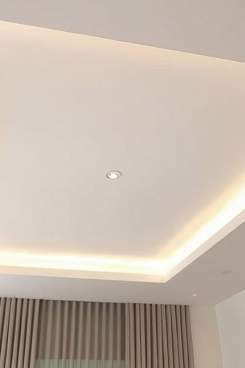 Plafond blanc avec éclairage encastré et bandeau lumineux périphérique. Rideaux beiges en dessous.