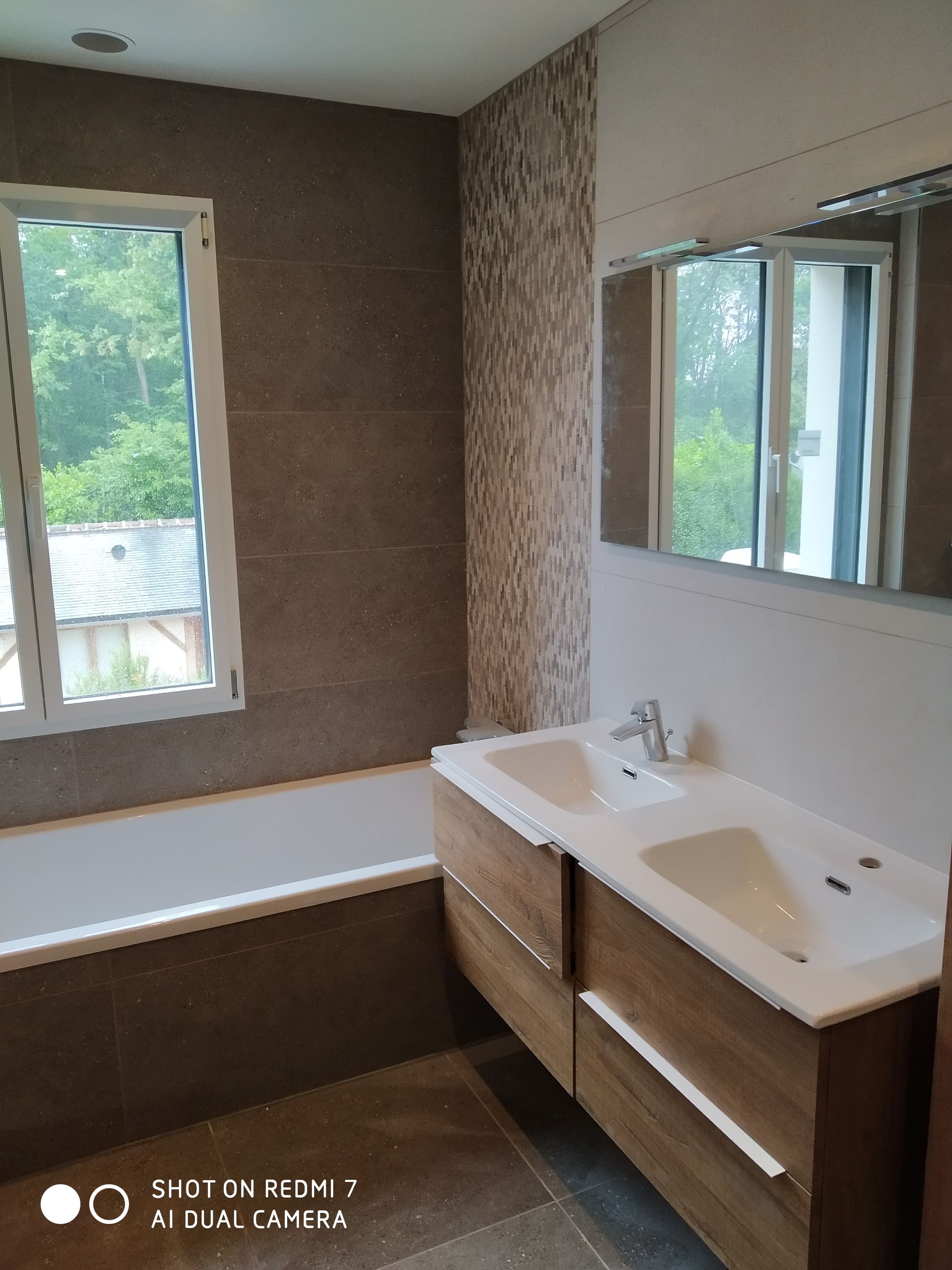 Salle de bains avec baignoire, meuble-lavabo en bois avec double vasque et fenêtre donnant sur la verdure.